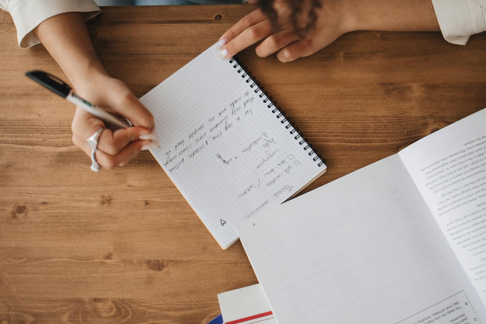 Close-up view of a person taking notes in a notebook with an open book on a wooden desk — UPI Study