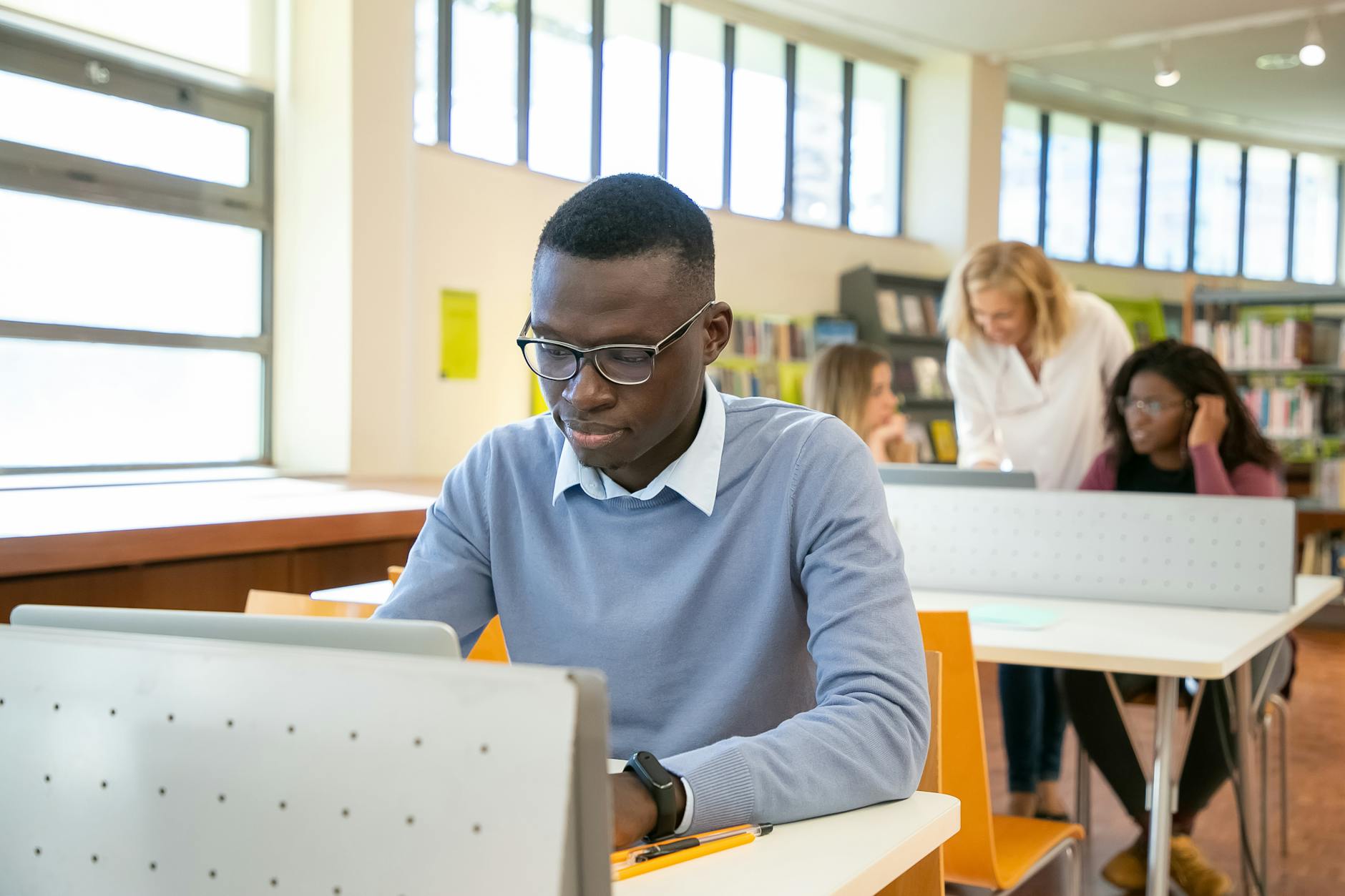 Concentrated young black male student in casual clothes and eyeglasses using laptop sitting at desk in classroom with multiethnic group of mates in college — UPI Study