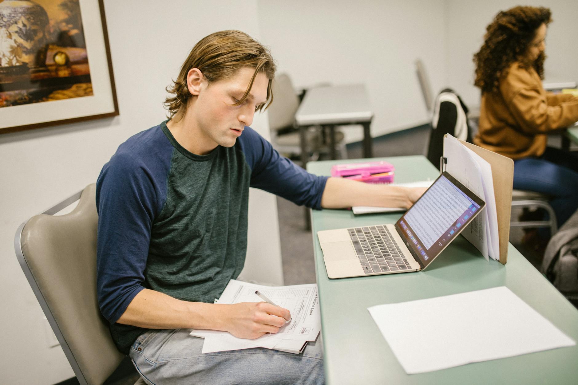 College student writing notes and using a laptop at a classroom desk for exam preparation — UPI Study