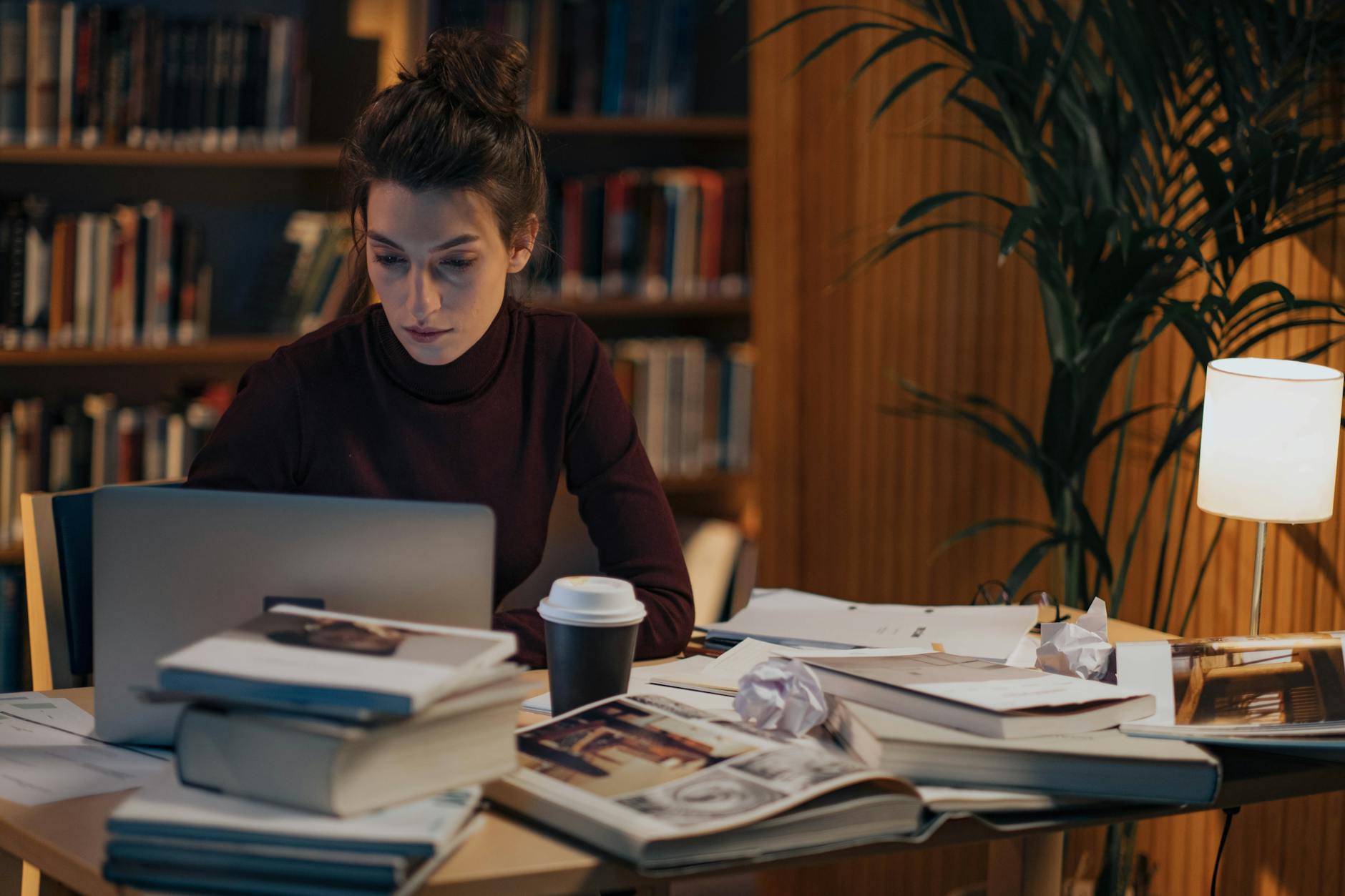 Woman working late at a desk filled with books, laptop, and coffee in a cozy library setting — UPI Study
