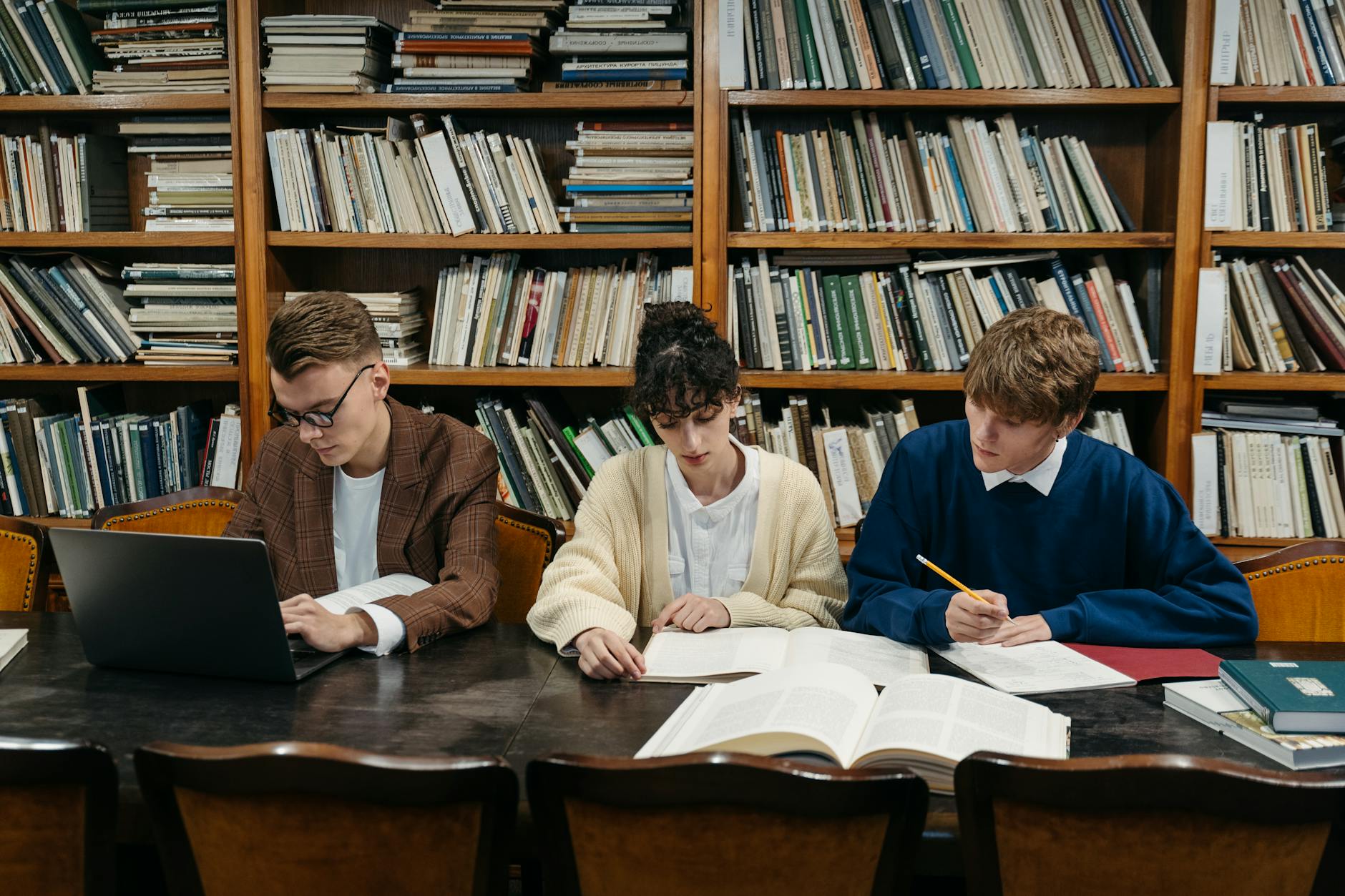 Three students work on laptops and books in a university library filled with shelves — UPI Study