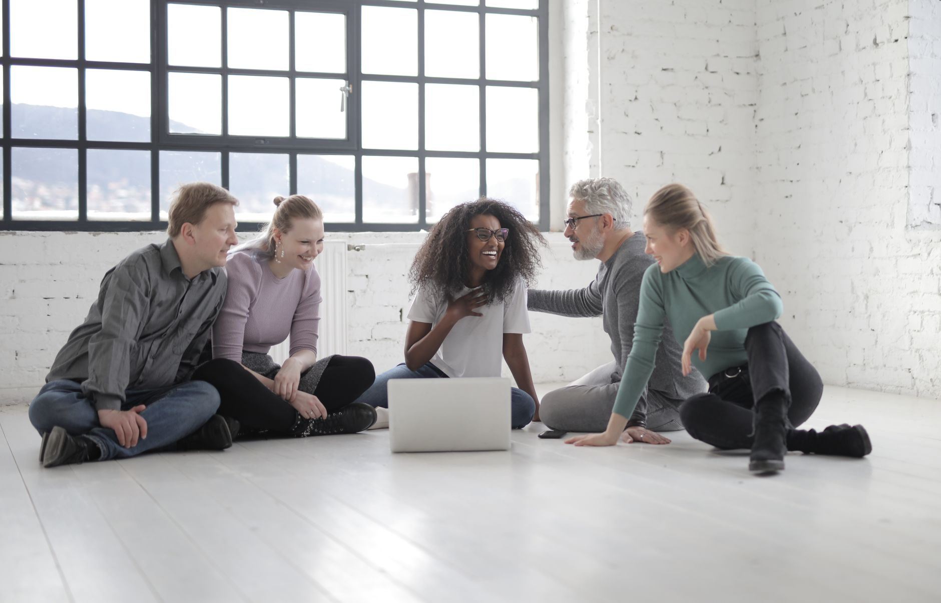 Happy multiethnic group of people gathering together while speaking and sitting with crossed legs in front of portable computer on ceramic floor near window in sunlight — UPI Study