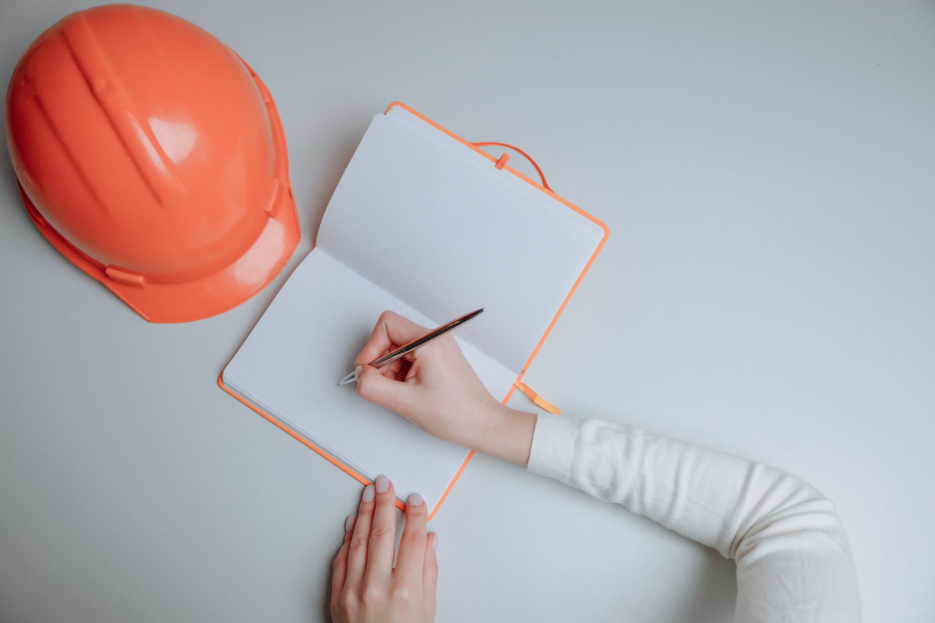 A top view of a person writing in a notebook next to an orange hardhat on a white background — UPI Study