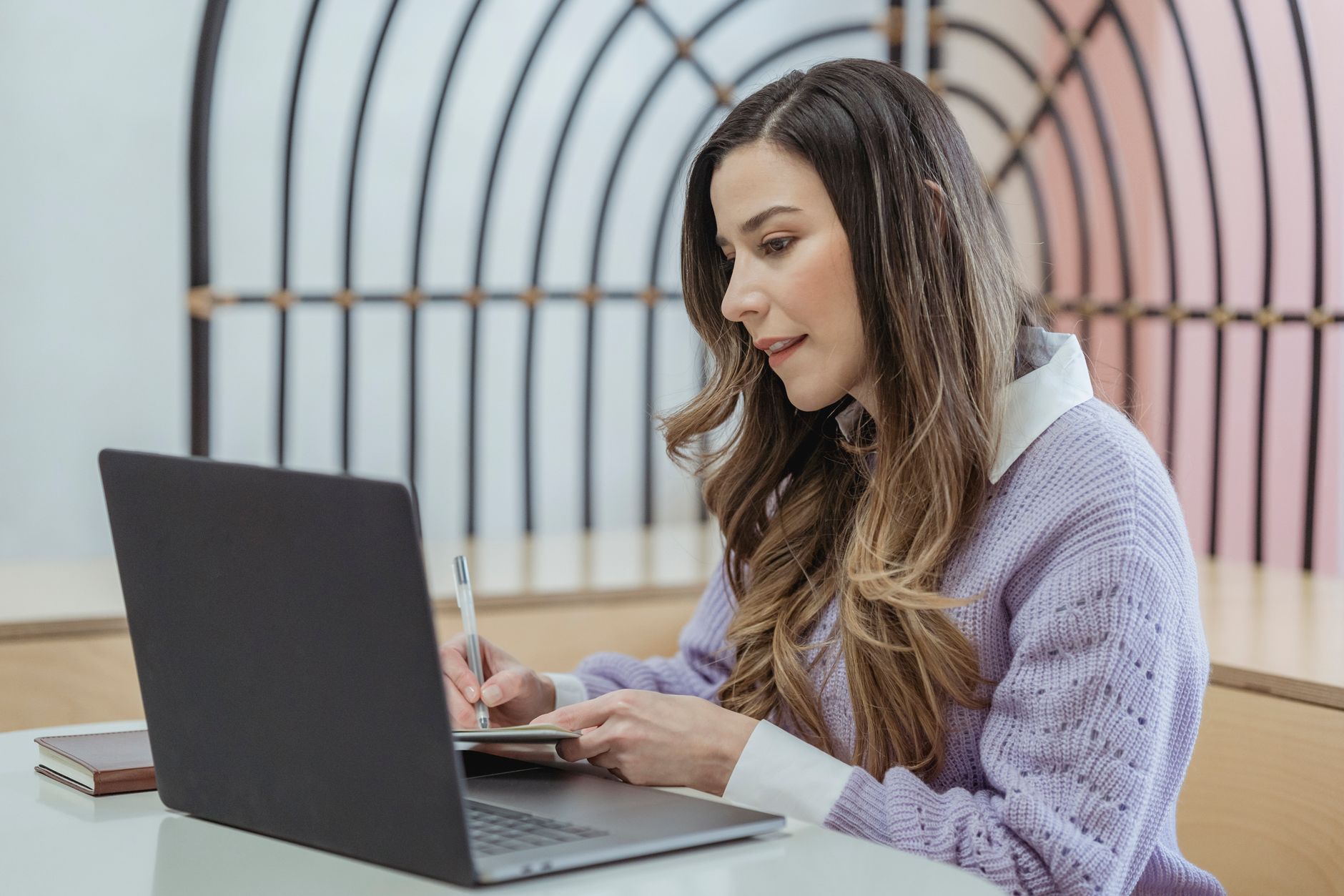 Focused female freelancer with wavy hair writing notes in planner while sitting at table and working on netbook — UPI Study