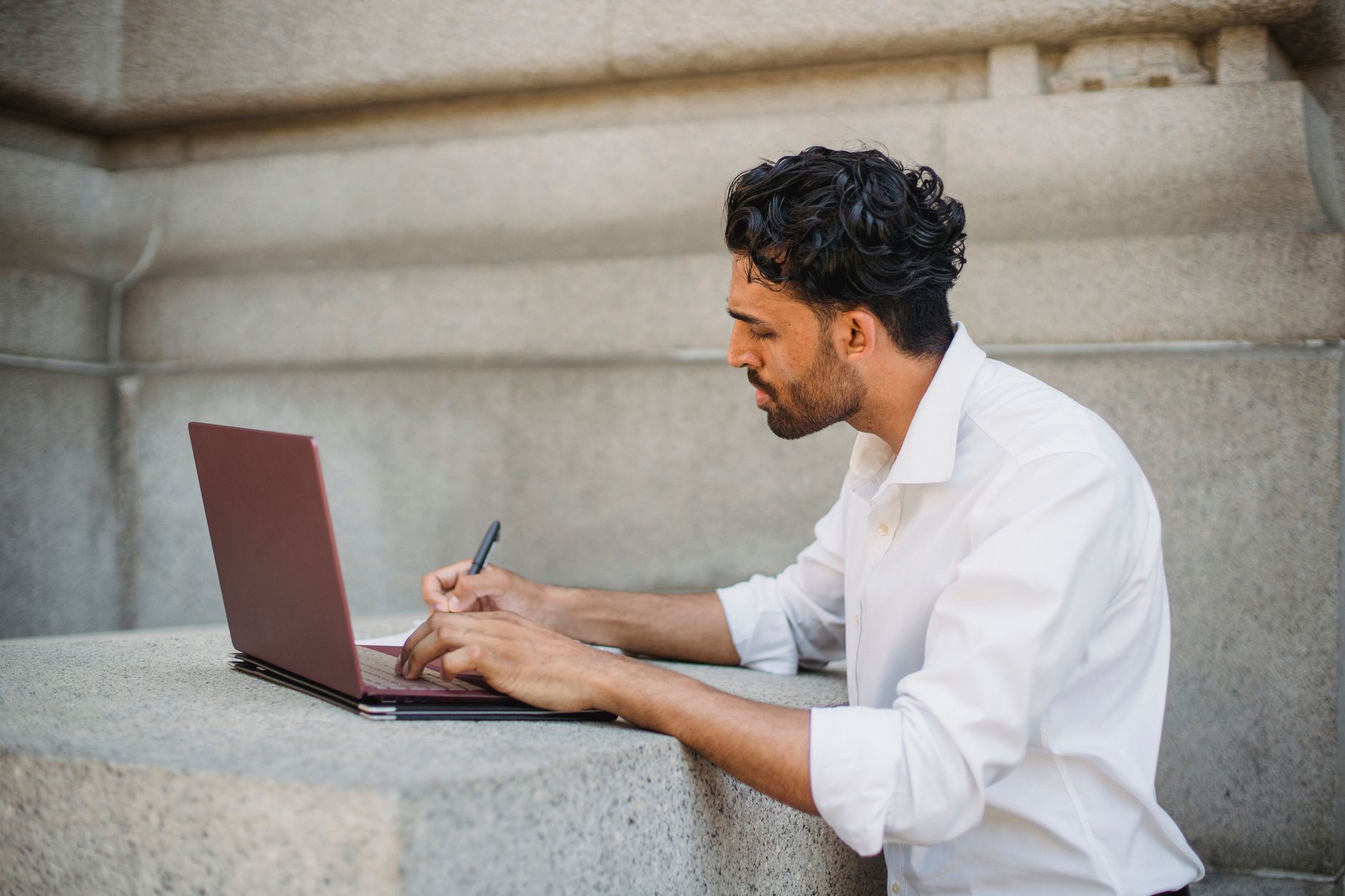 A man writing on a laptop outdoors, seated on a stone surface and focused on work — UPI Study
