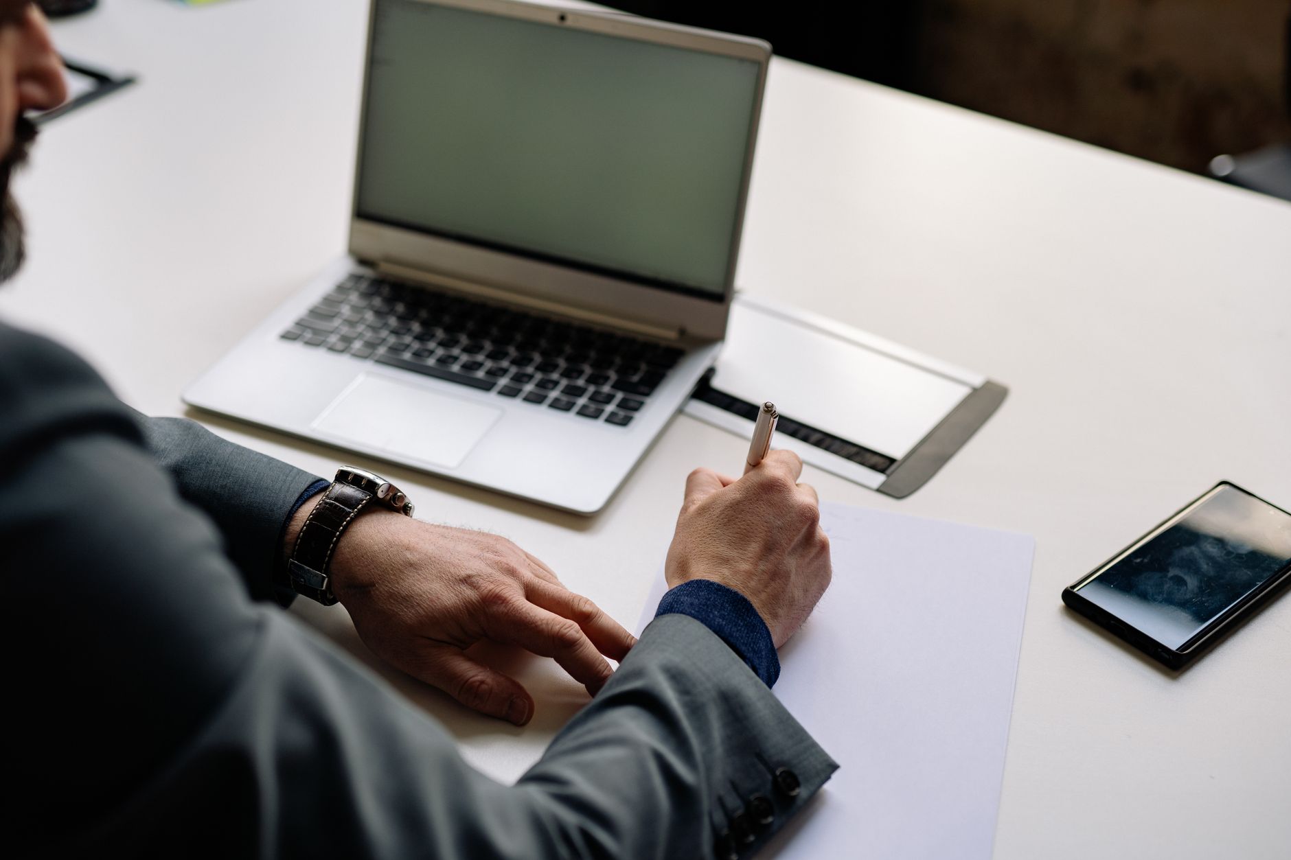 Professional businessman writing notes in office setting with laptop and smartphone visible — UPI Study
