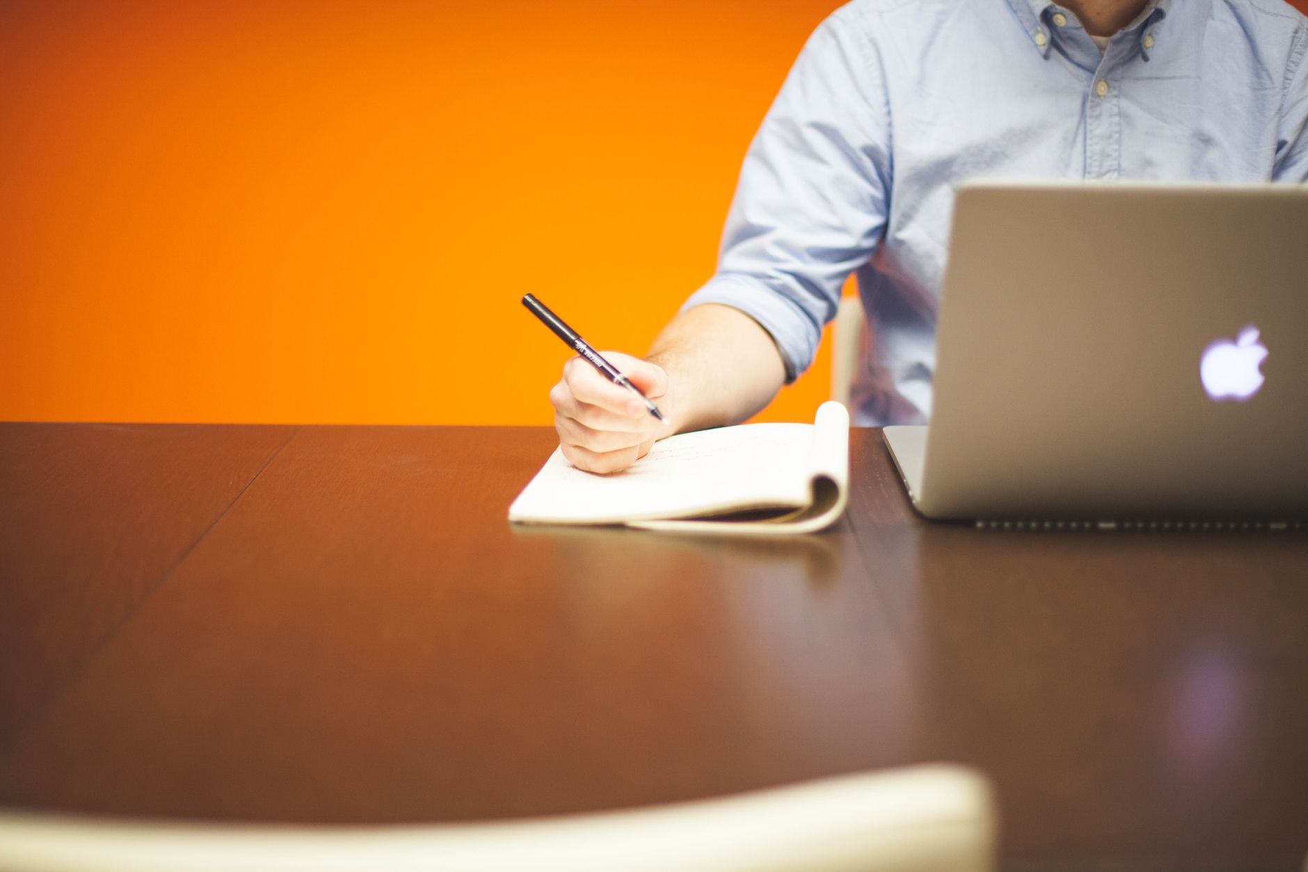 Adult writing notes in a notebook beside a laptop on a wooden desk with orange background — UPI Study