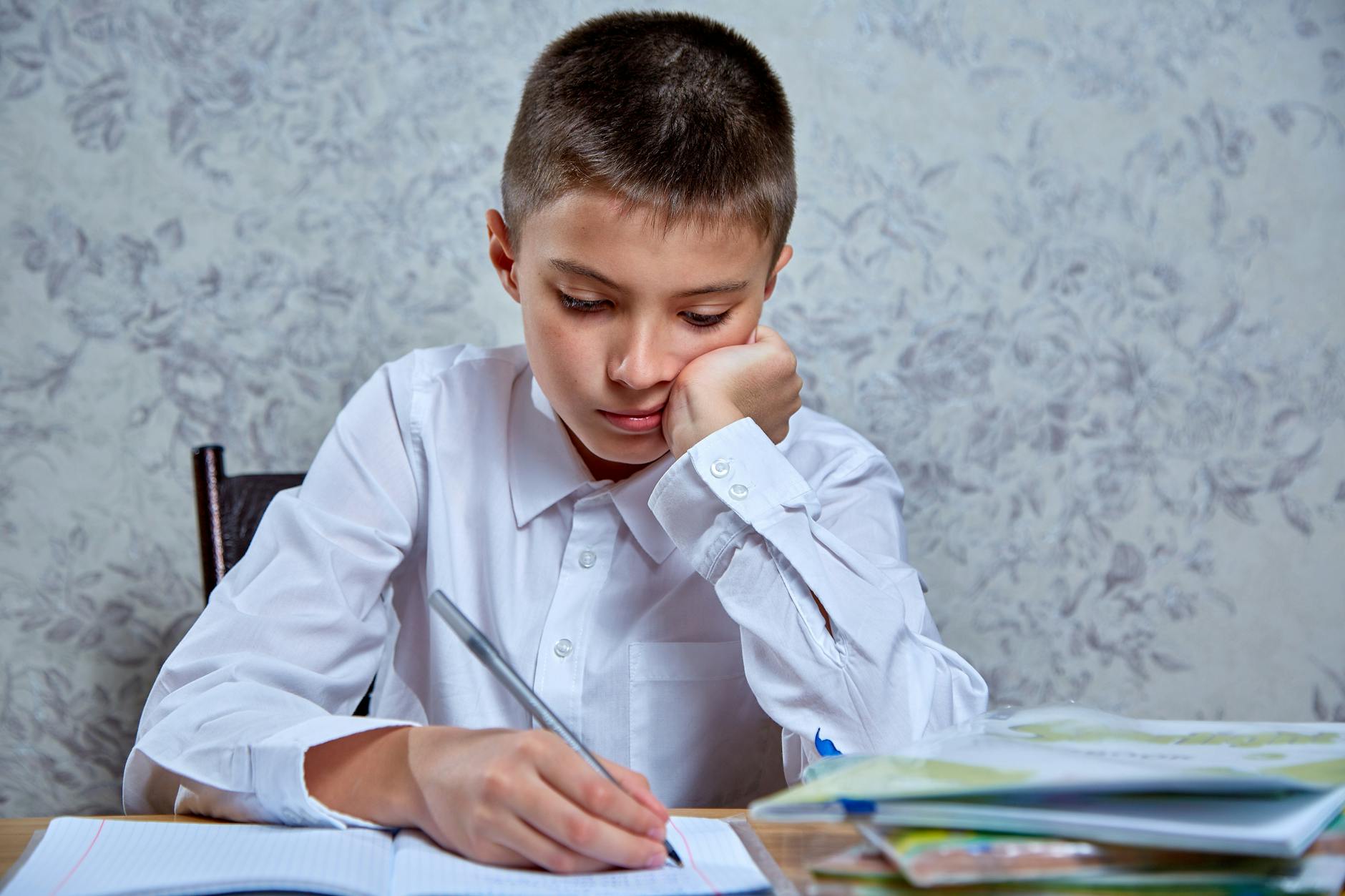 A young boy in a white shirt attentively writing in a notebook at a desk, indoors — UPI Study