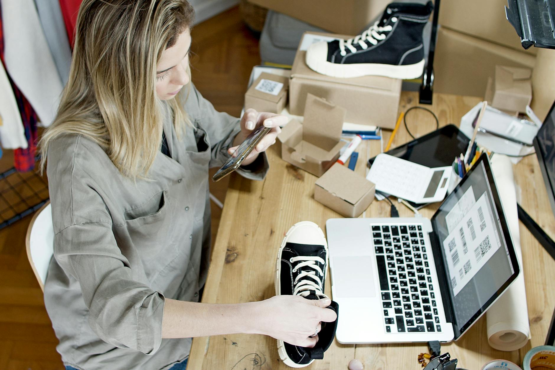 Woman photographing shoes for online sale in home workspace with laptop and packages — UPI Study