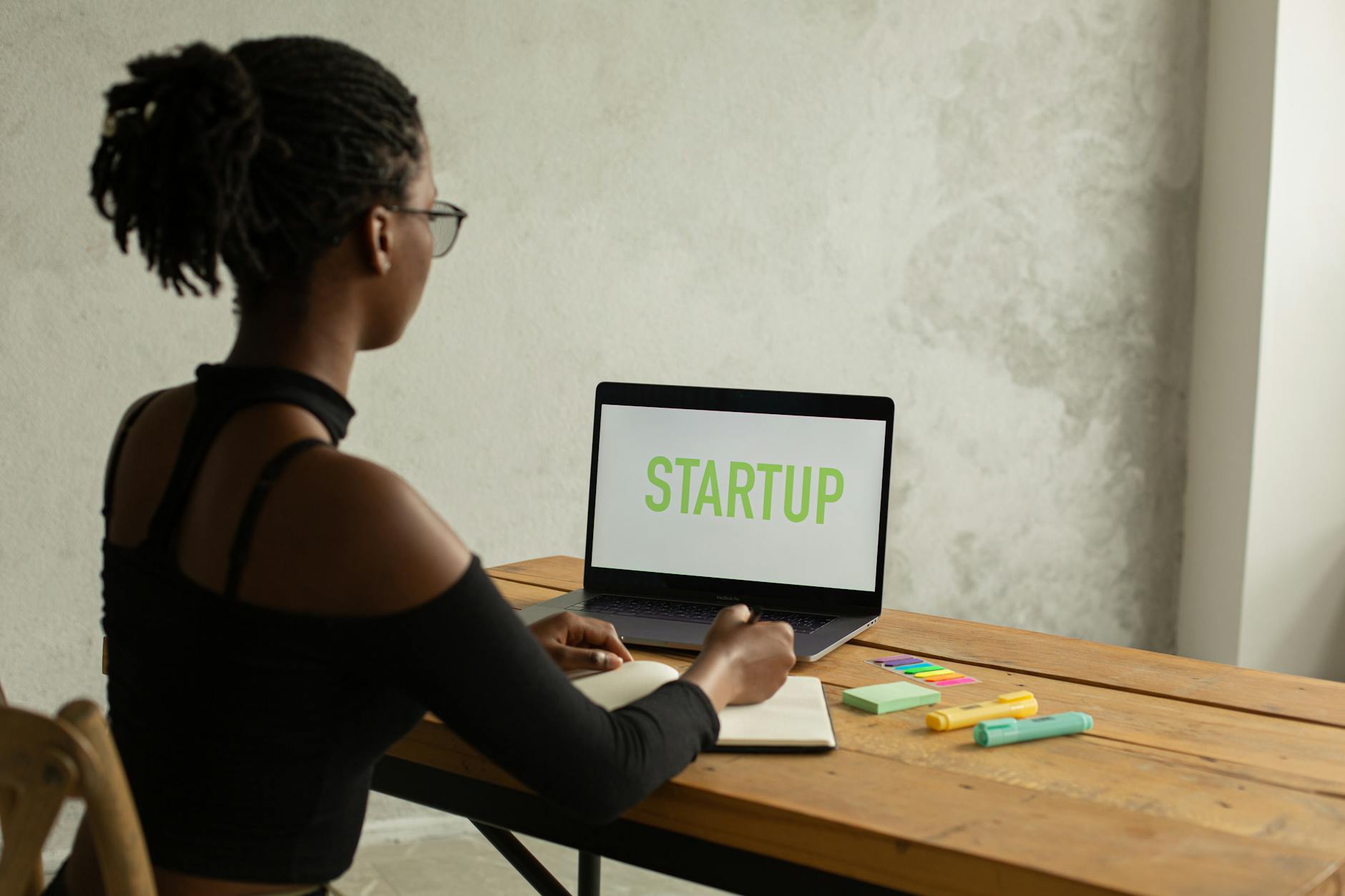 Back view of anonymous young African American businesswoman with bare shoulder and eyeglasses sitting at table with netbook with startup on screen and writing notes in copybook — UPI Study