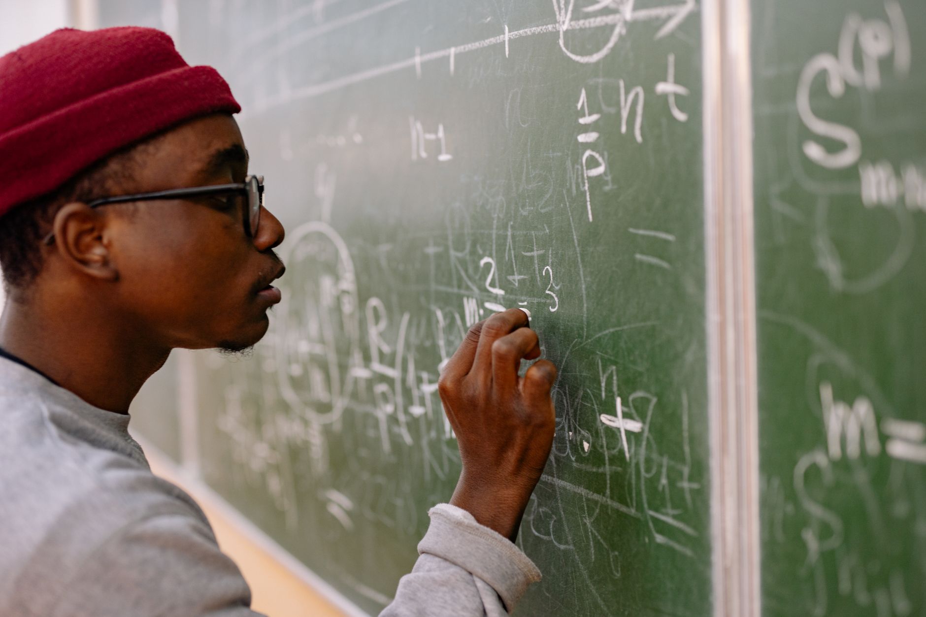 A young man with eyeglasses and a red beanie solving math problems on a chalkboard — UPI Study