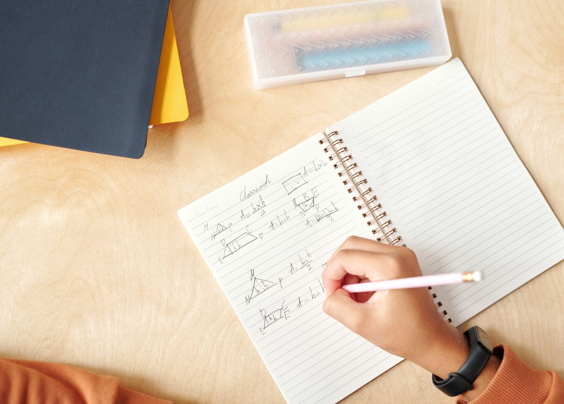 A student writes geometric formulas by hand in a notebook on a desk — UPI Study