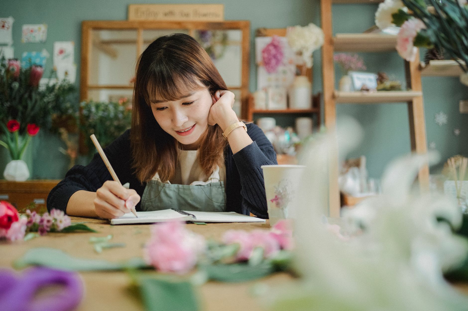 Smiling young female florist in apron writing notes while working in cozy flower shop — UPI Study
