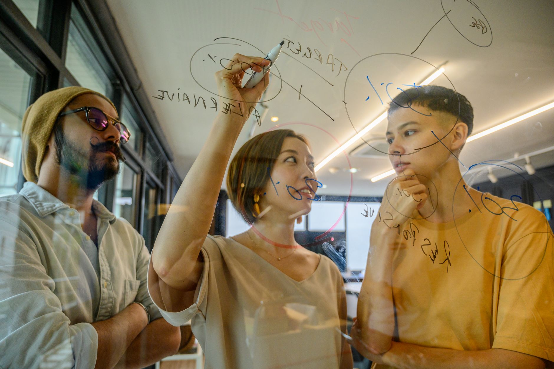 Three diverse young adults collaborating and brainstorming ideas on a glass board in a modern office setting — UPI Study