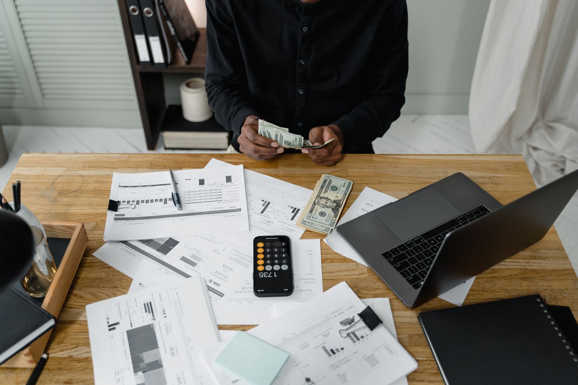 A person counts dollar bills at a desk with a laptop, papers, and calculator — UPI Study