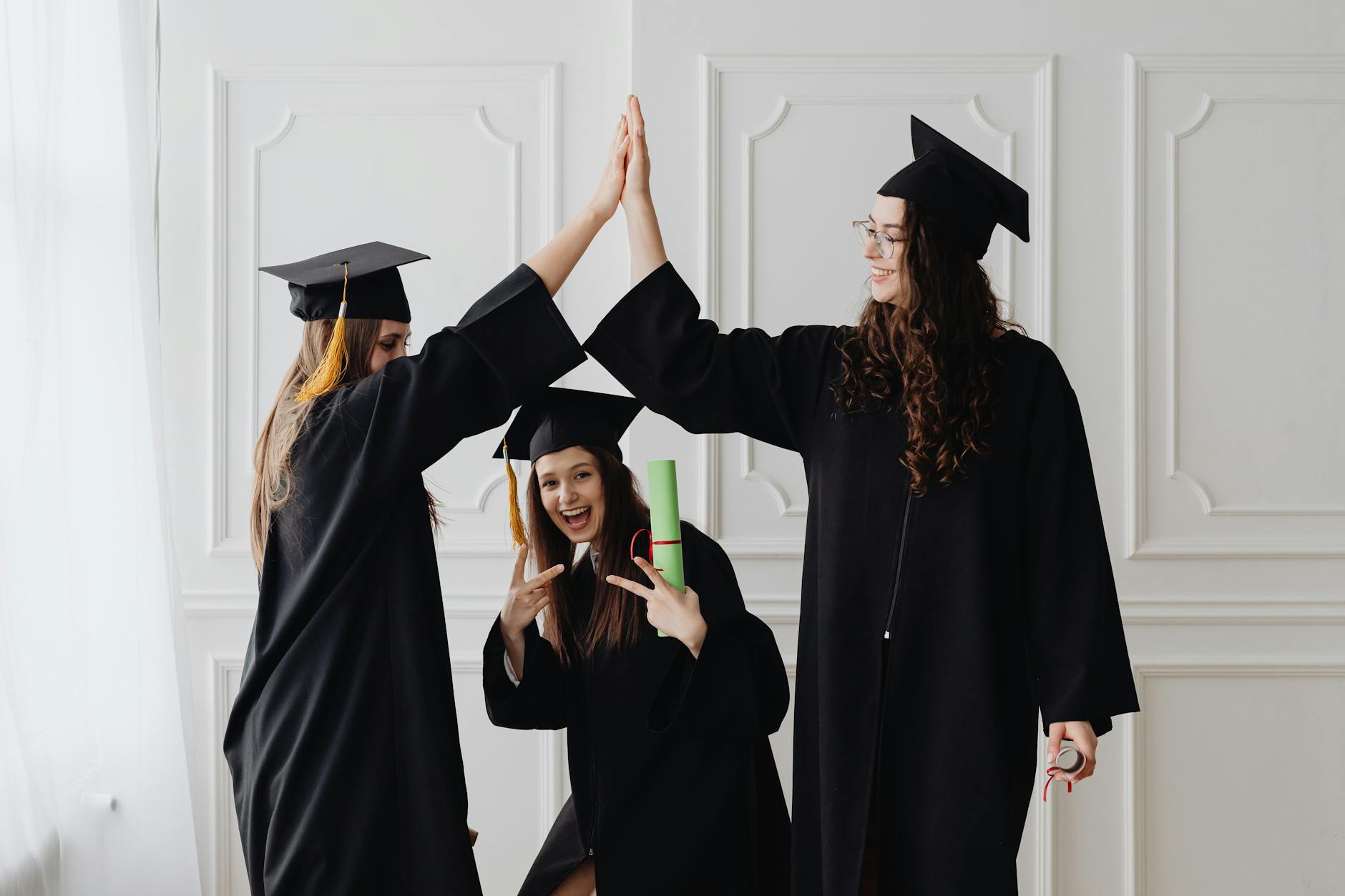 Three young women celebrating graduation with a high five, holding diplomas indoors — UPI Study