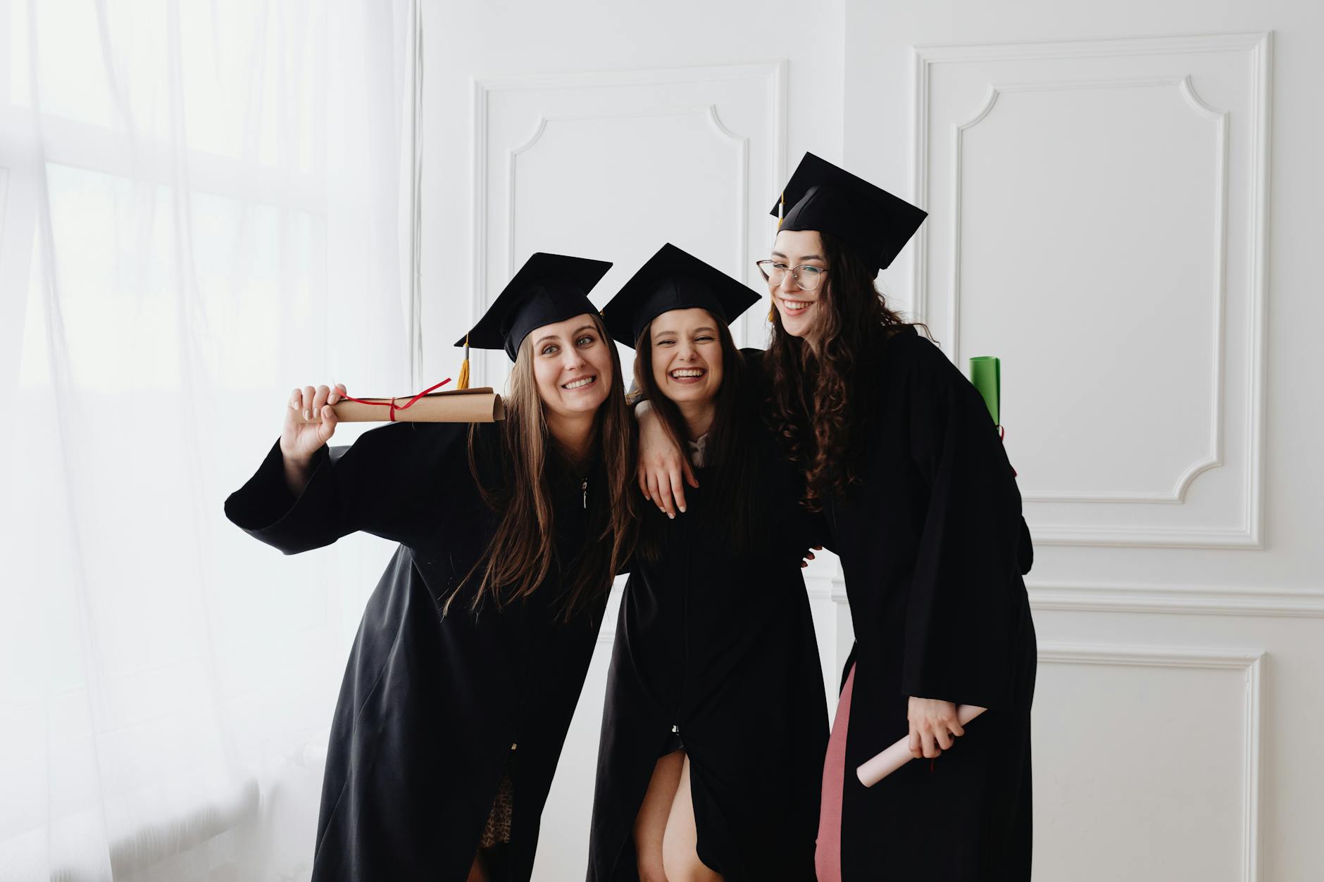 Three smiling graduates in caps and gowns celebrating indoors together — UPI Study