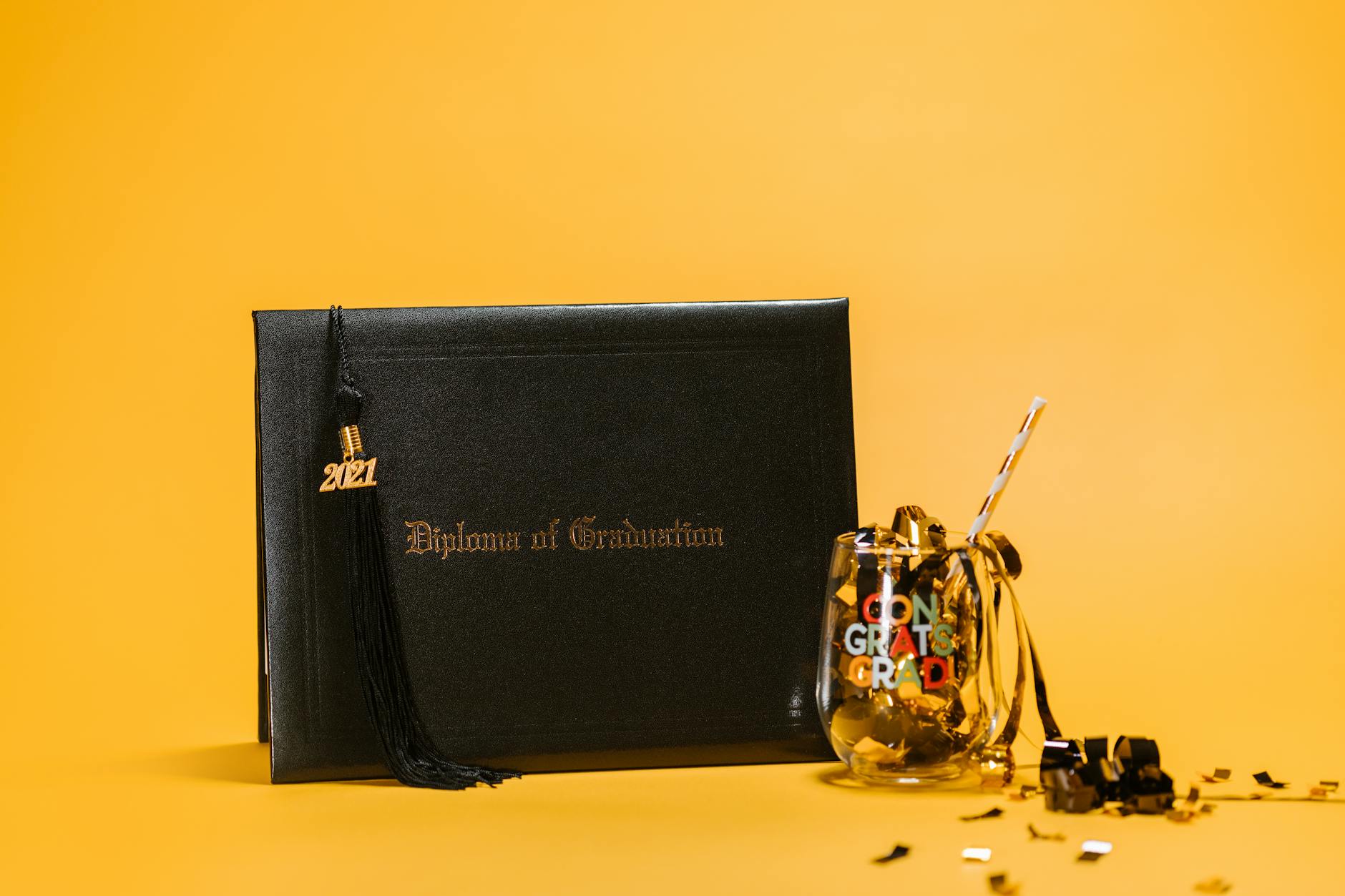 Black diploma and glass with confetti against a yellow background, symbolizing graduation celebration — UPI Study