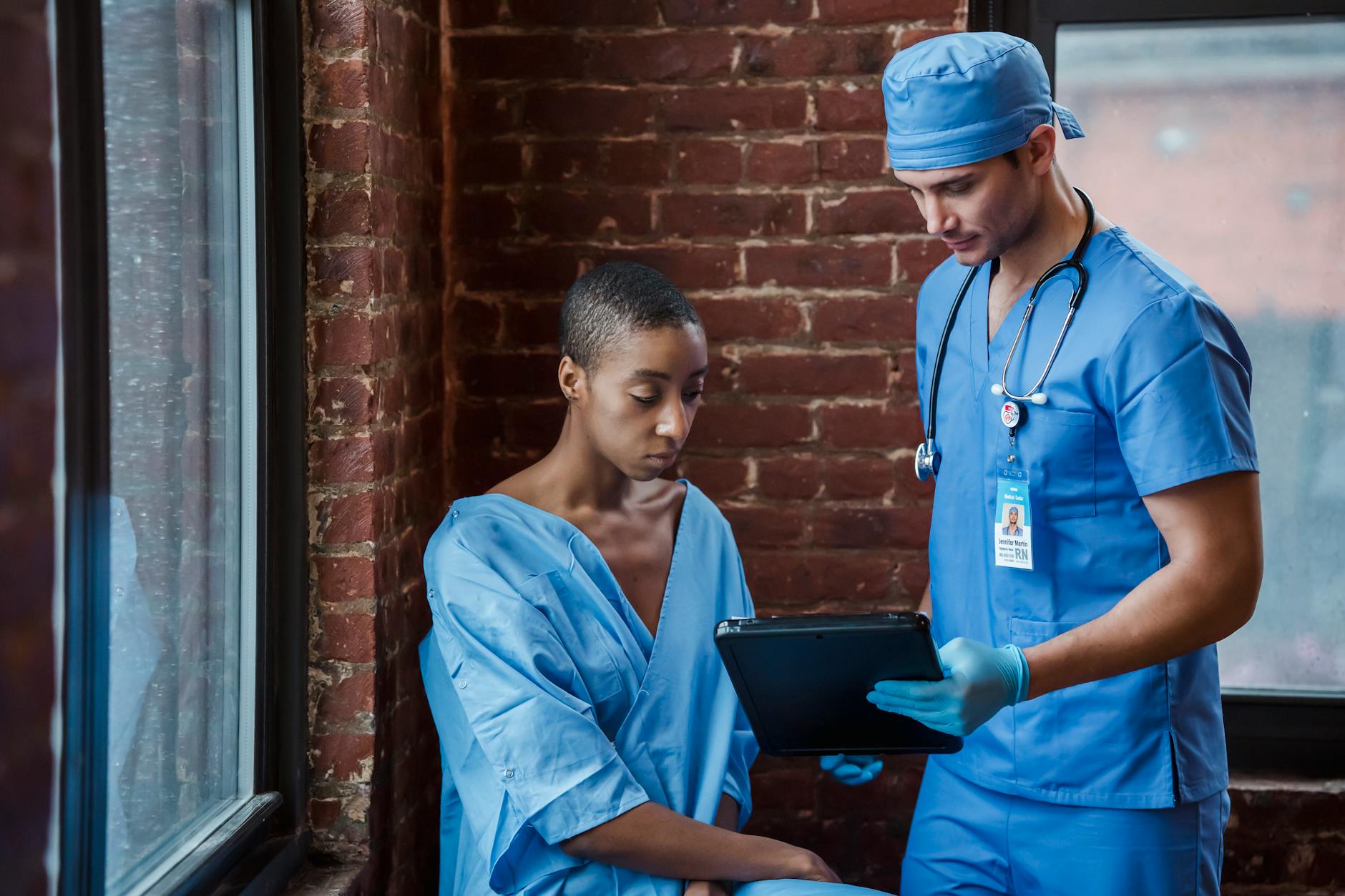 Thoughtful doctor in uniform and gloves showing diagnosis to African American female patient with short hair in blue medical robe in hallway of hospital in daytime — UPI Study