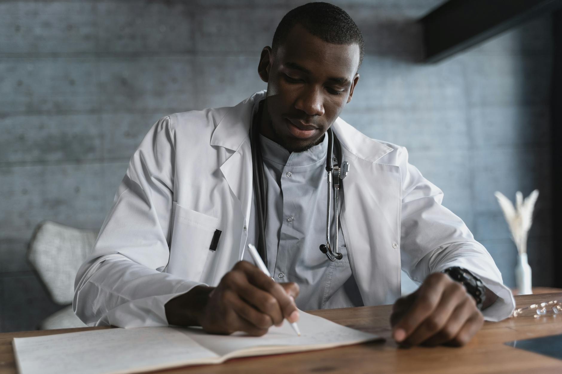 Black male doctor in a lab coat writing notes at a wooden desk in a medical office — UPI Study