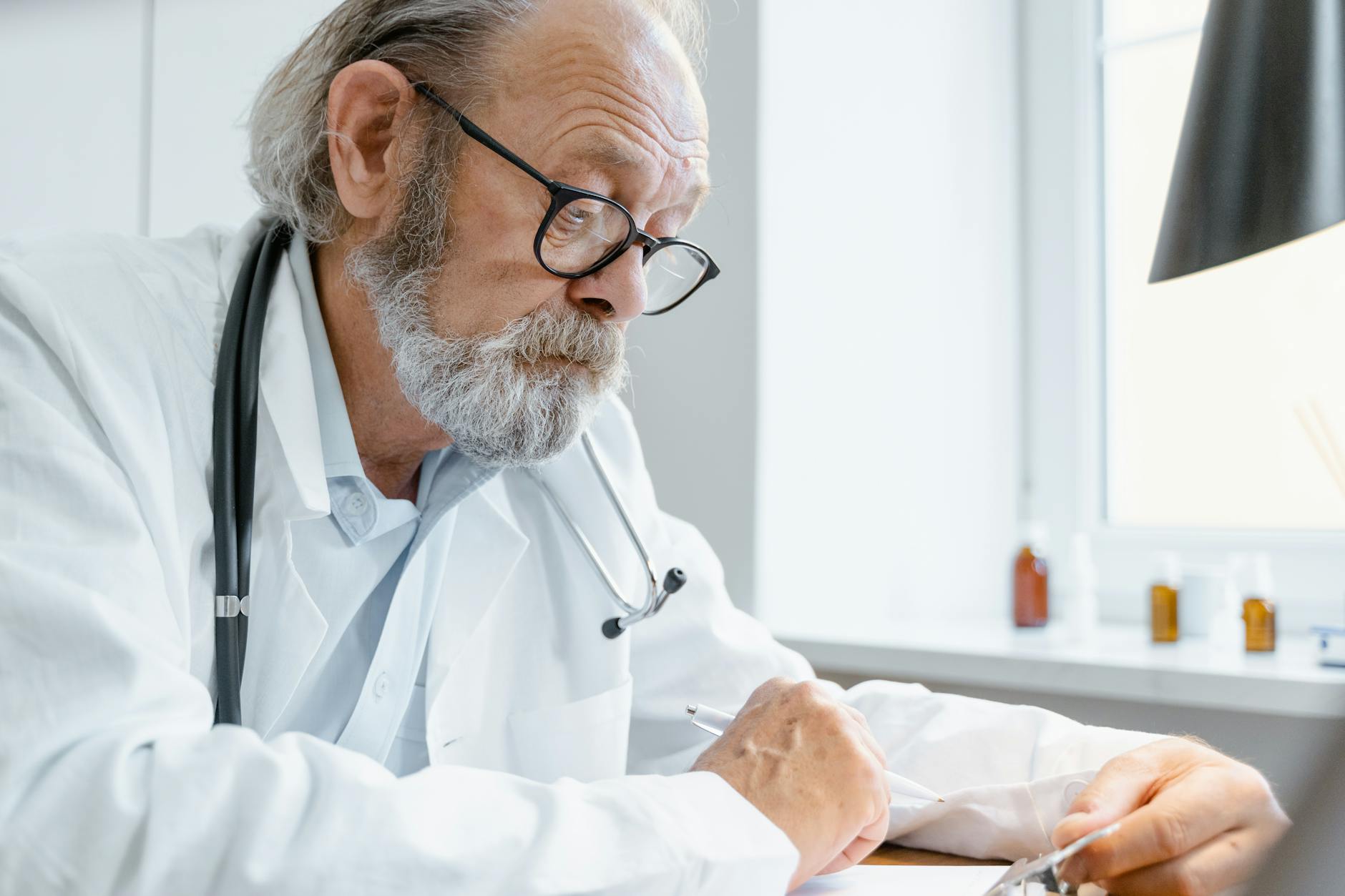 Elderly doctor wearing eyeglasses and stethoscope writes at office desk — UPI Study