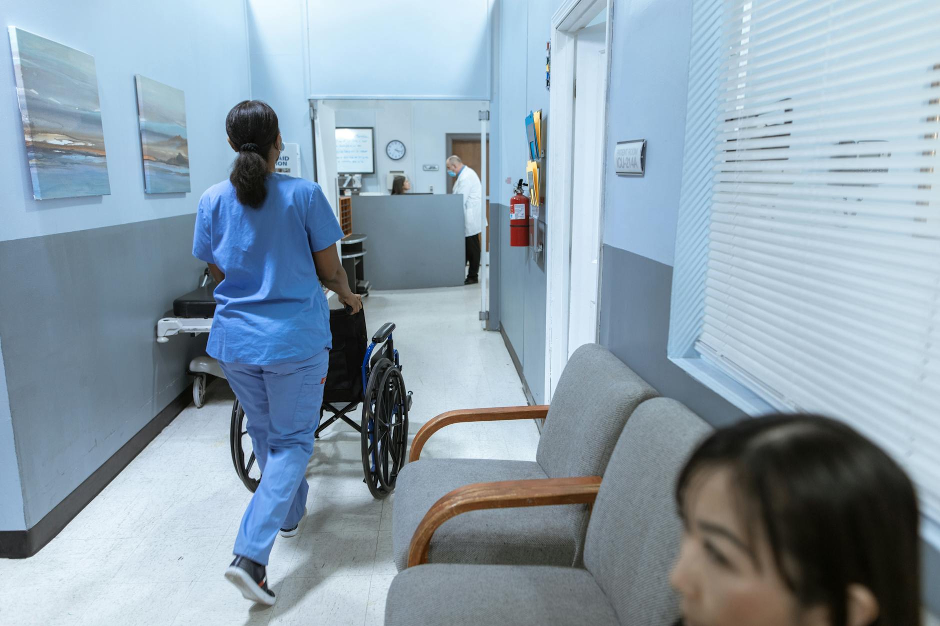 A nurse in scrubs pushes a wheelchair through a well-lit hospital corridor, attending to patient care — UPI Study