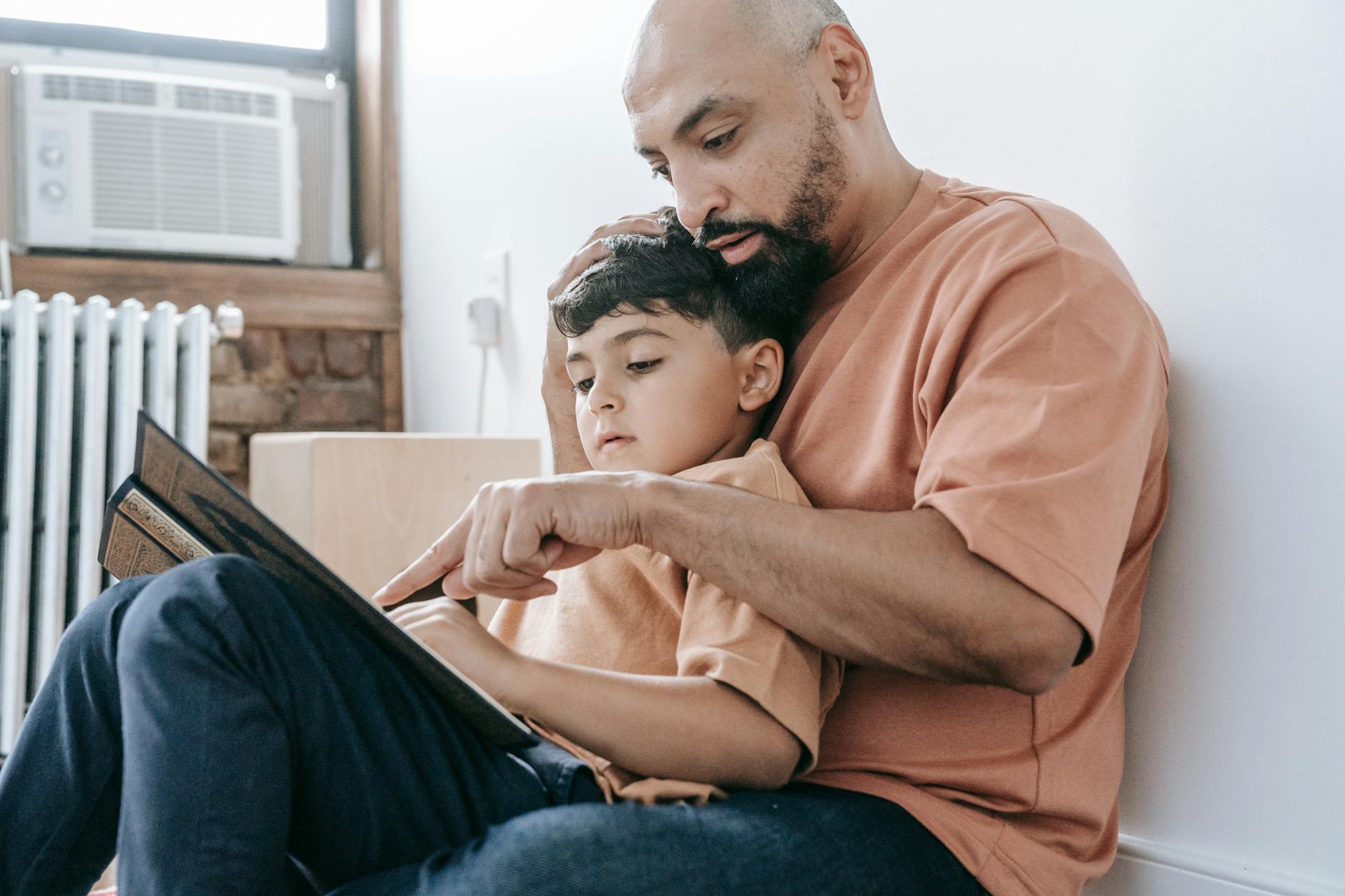 A father in an orange shirt reads a book to his son while sitting indoors, fostering learning and bonding — UPI Study