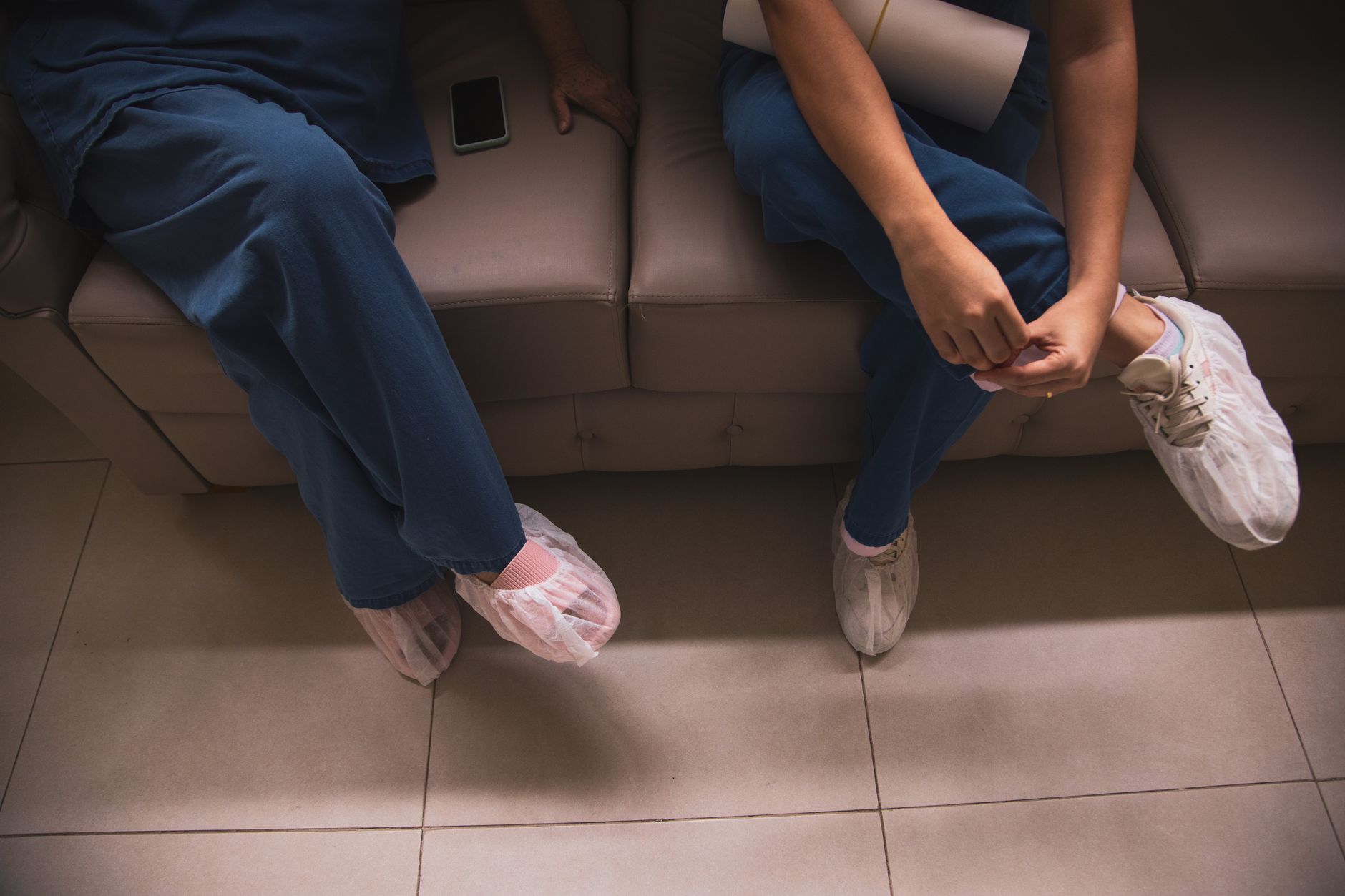 Two healthcare workers sitting on a sofa wearing scrubs and protective shoe covers — UPI Study