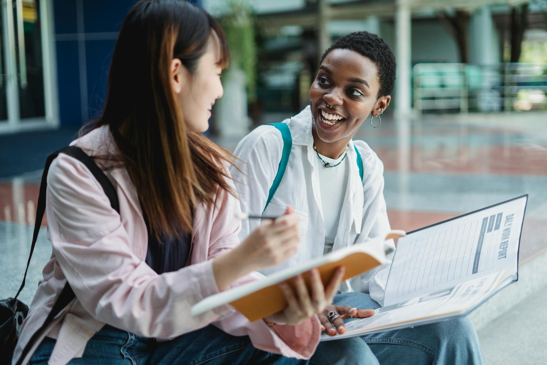 Happy young multiracial women in casual clothes sitting outdoors with notepad and papers in daytime while looking at each other — UPI Study
