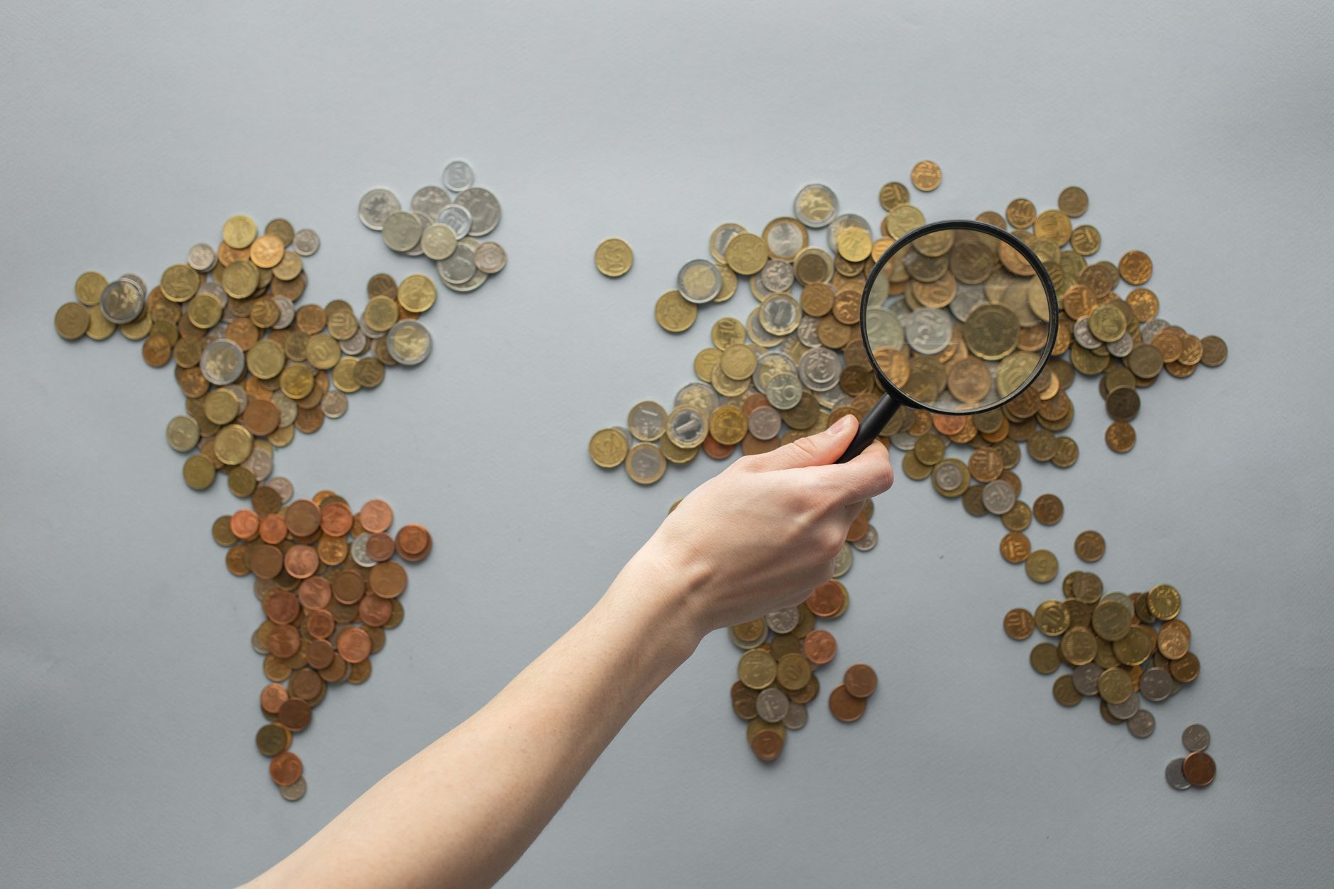 Top view of crop unrecognizable traveler with magnifying glass standing over world map made of various coins on gray background — UPI Study