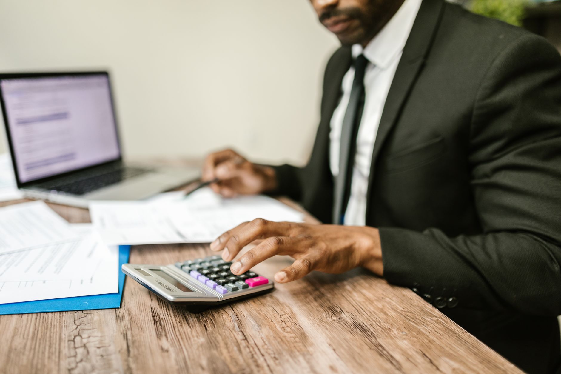 A businessman in a black suit using a calculator at a desk with financial documents and a laptop — UPI Study