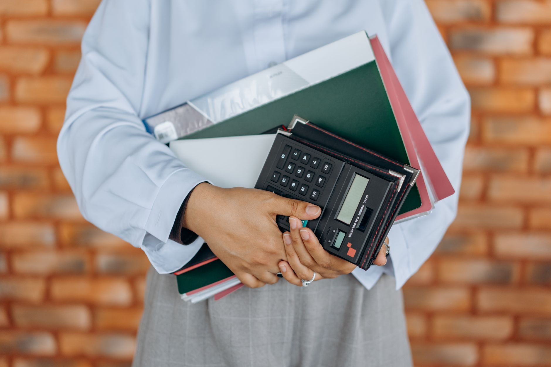 Close-up of a person holding a calculator and folders against a brick wall background — UPI Study
