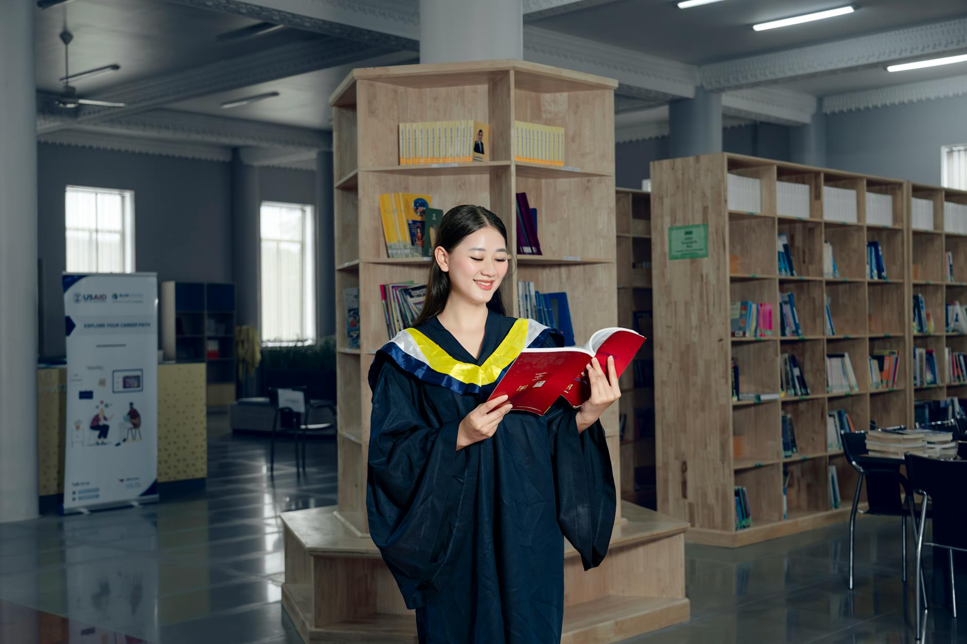 Woman in graduation gown reading a book in university library, symbolizing education and knowledge — UPI Study