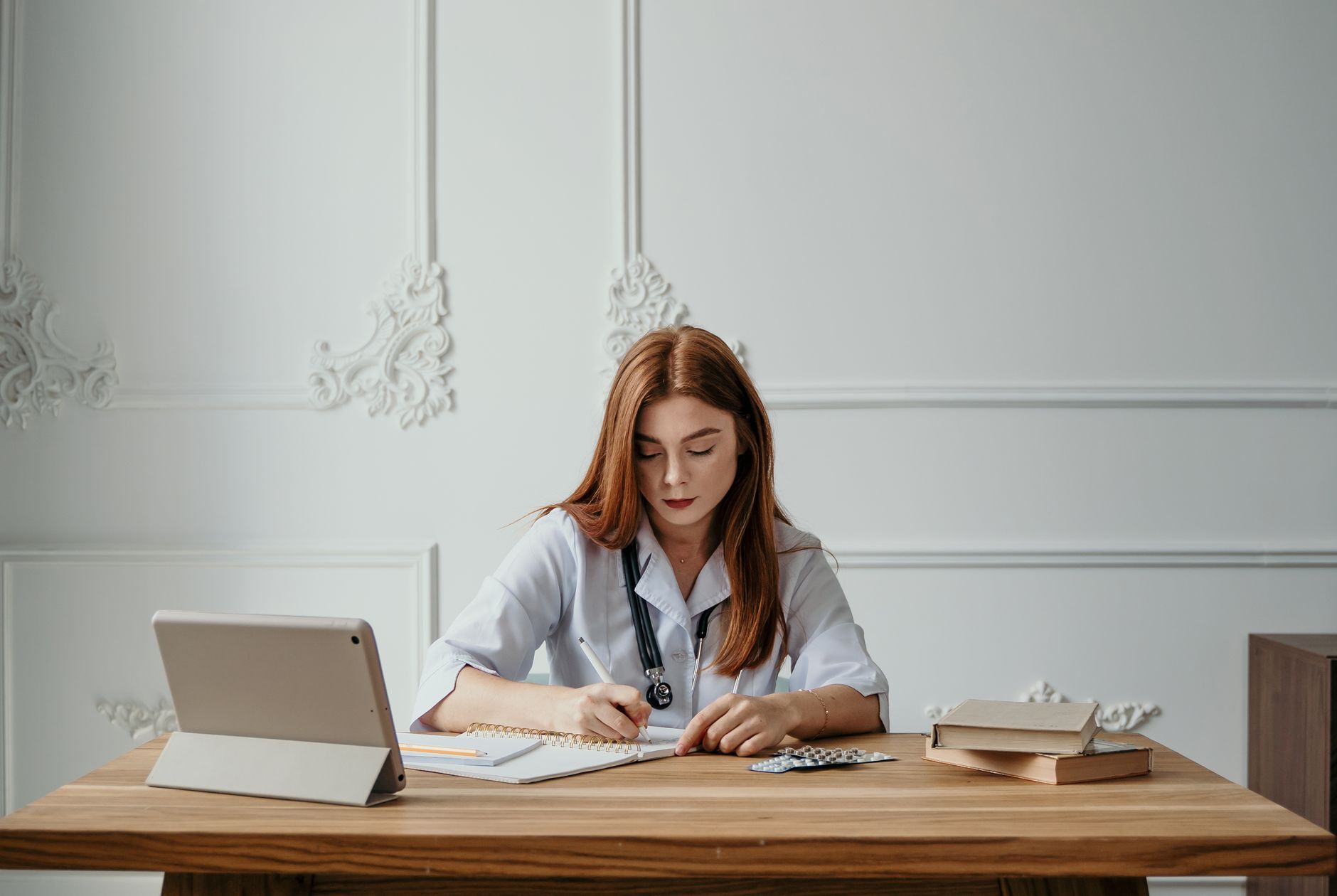 A woman doctor writing notes at a desk with a tablet and books, capturing a professional office setting — UPI Study