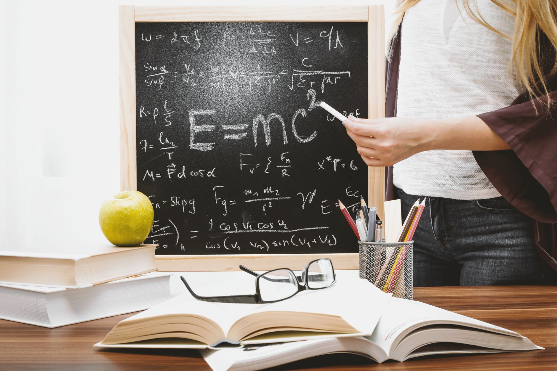 Woman writing physics equations on a blackboard with books and an apple on the desk — UPI Study