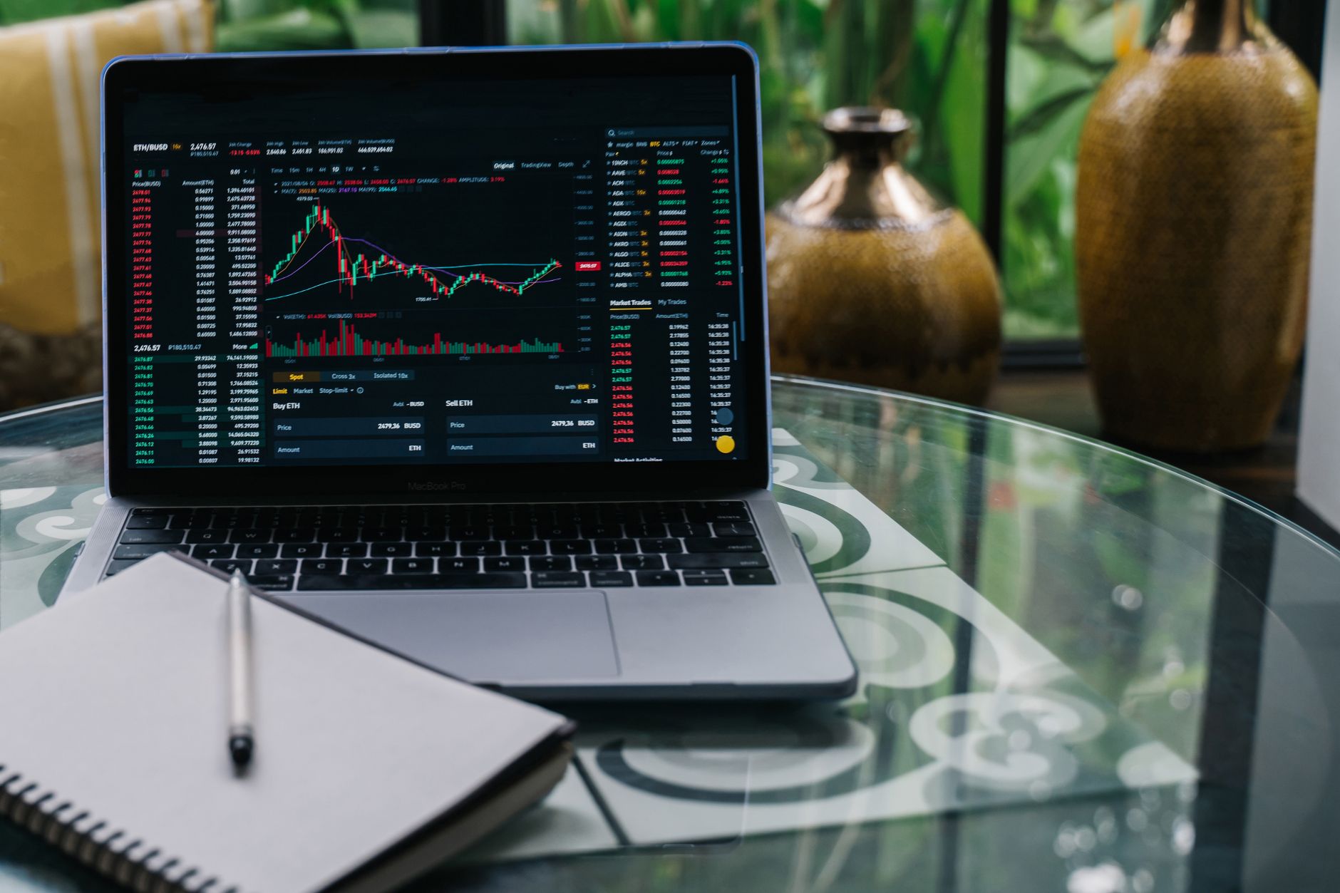 Laptop displaying cryptocurrency stocks and graphs on a glass table with a notepad — UPI Study