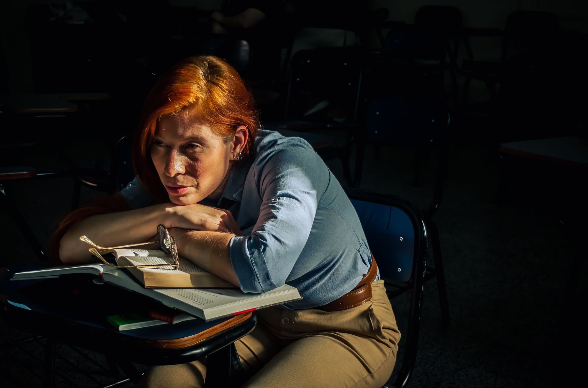 A student with red hair looks contemplative while resting on a desk in a dark classroom — UPI Study