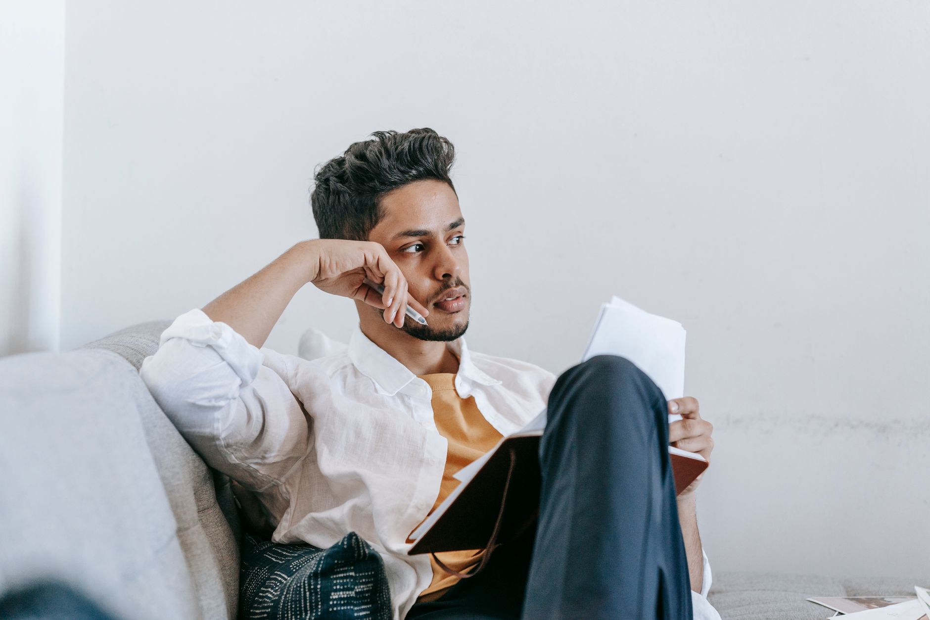 Contemplative young ethnic male in casual wear reading notes in planner and looking away in thoughts while resting on comfortable couch — UPI Study