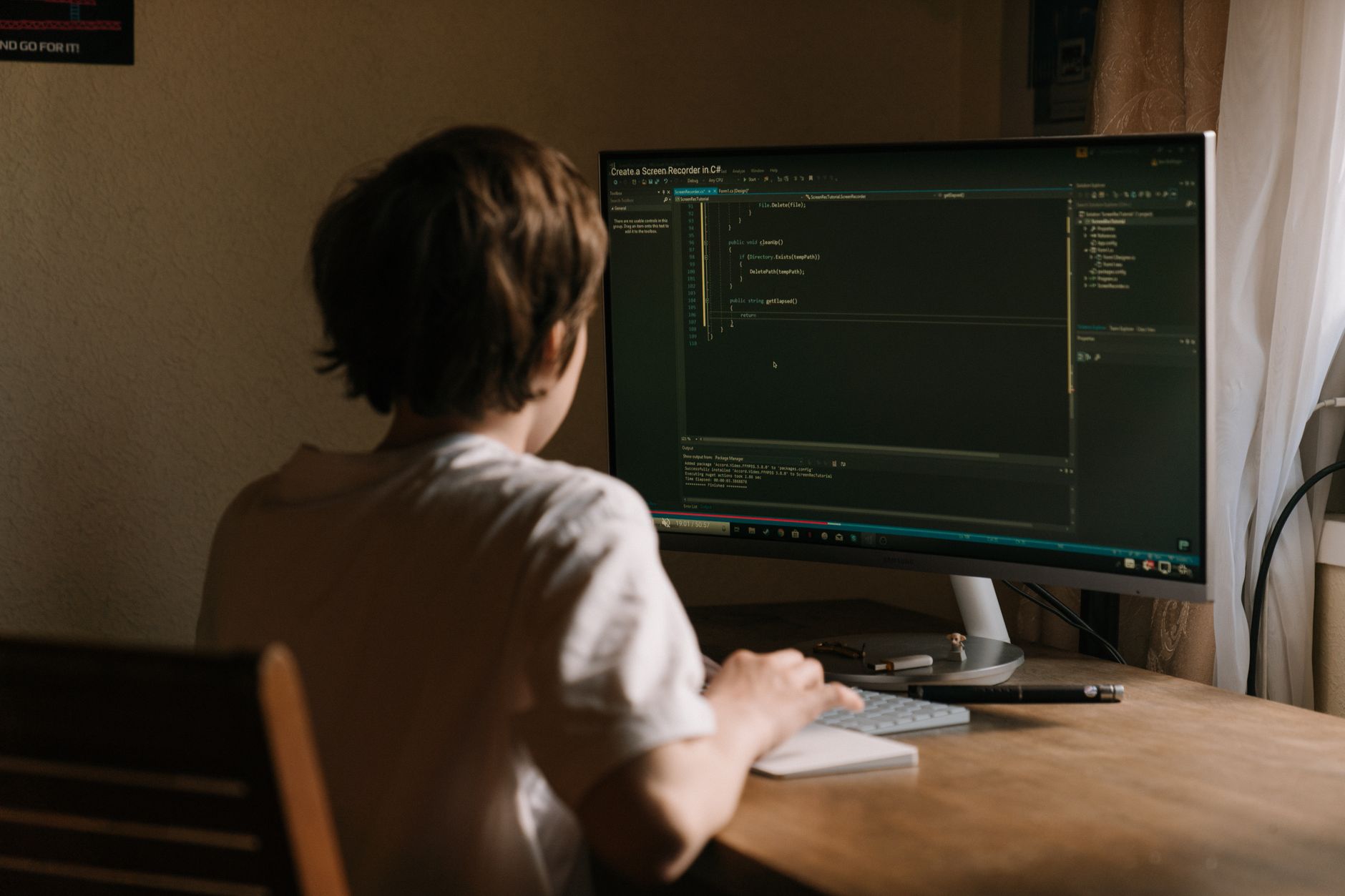 A teenager focused on coding software on a desktop monitor in a home office setting — UPI Study