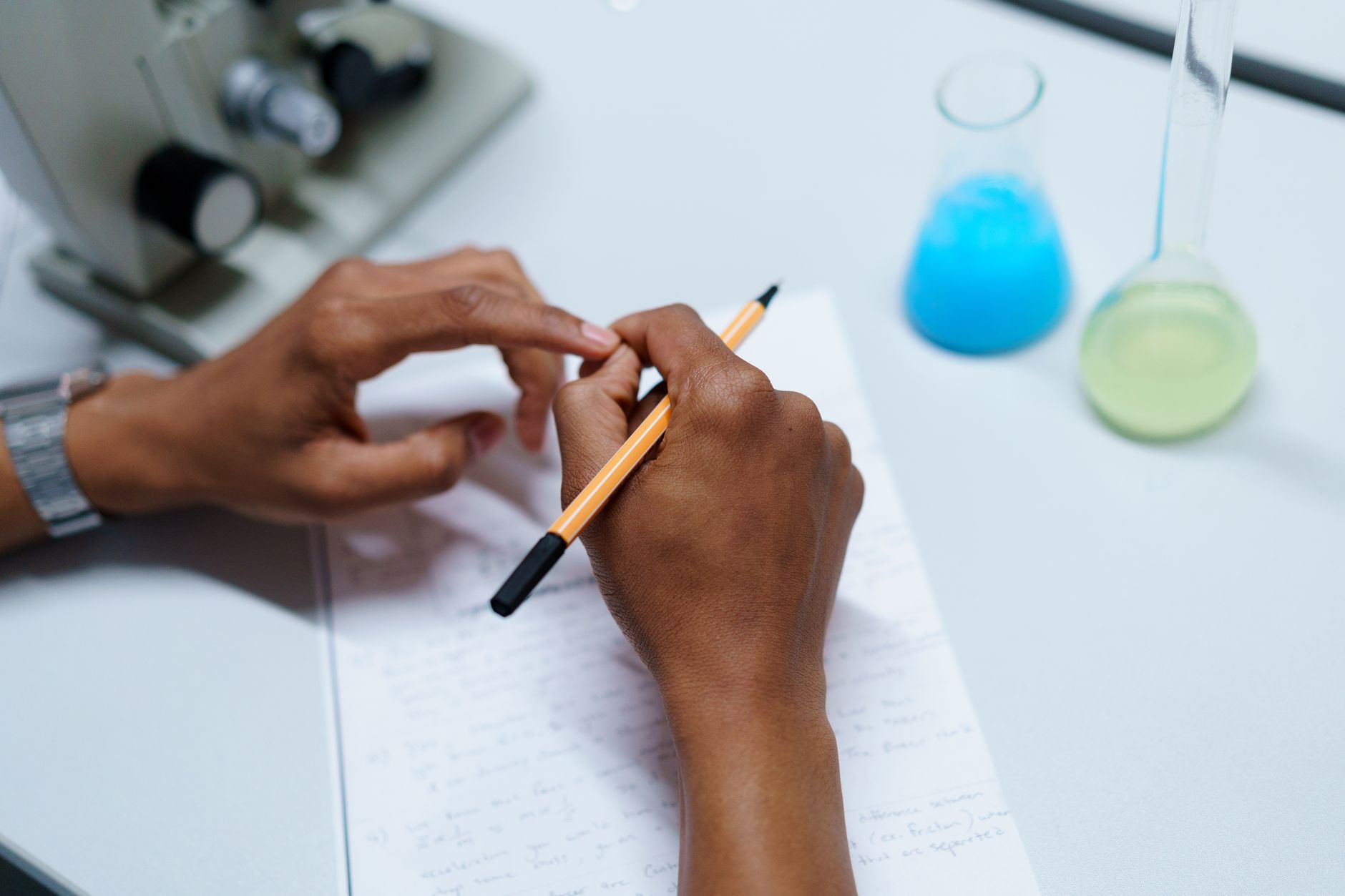Close-up of a scientist taking notes in a laboratory setting with flasks and a microscope — UPI Study