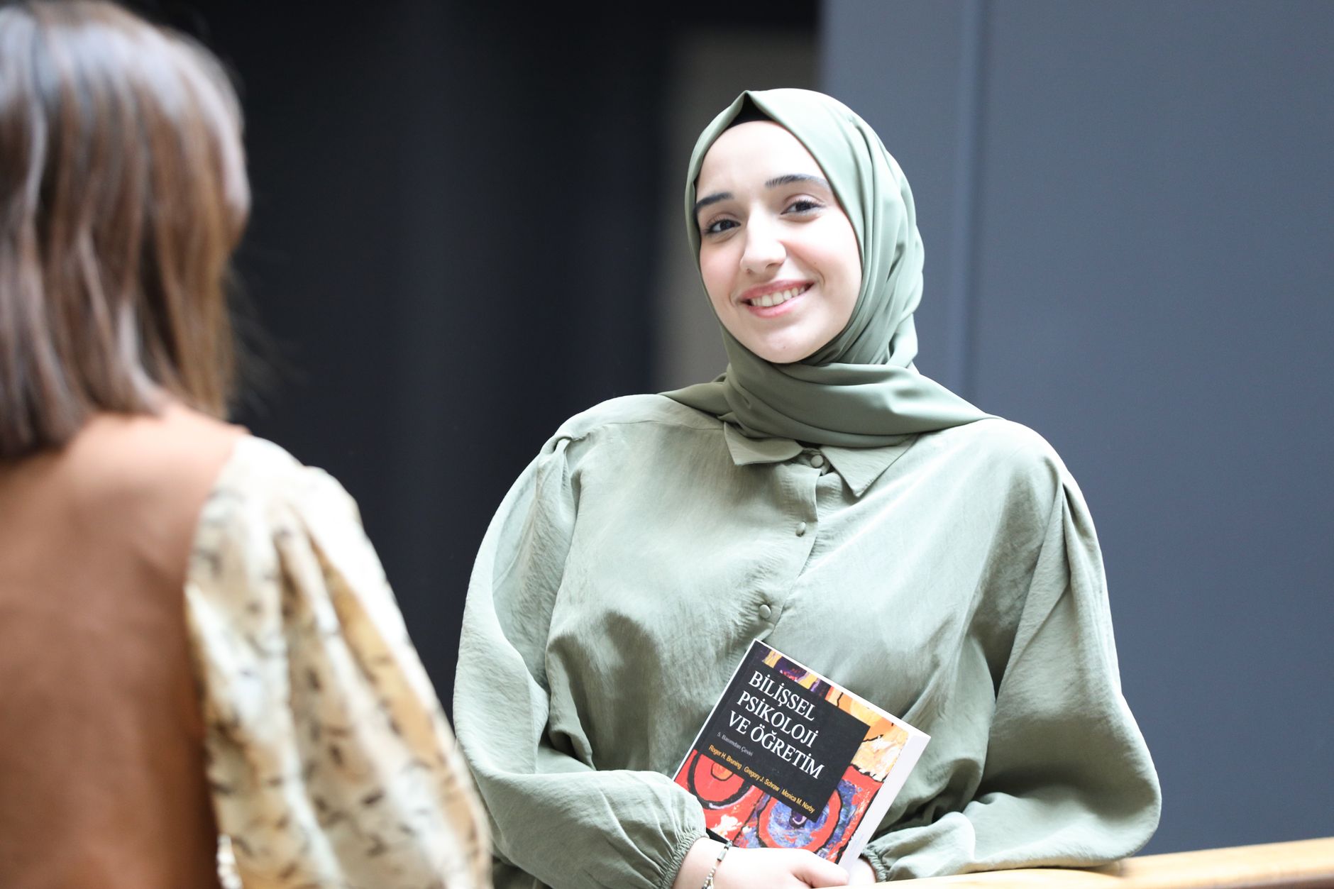A young woman in a headscarf smiles while holding a book, indoors — UPI Study
