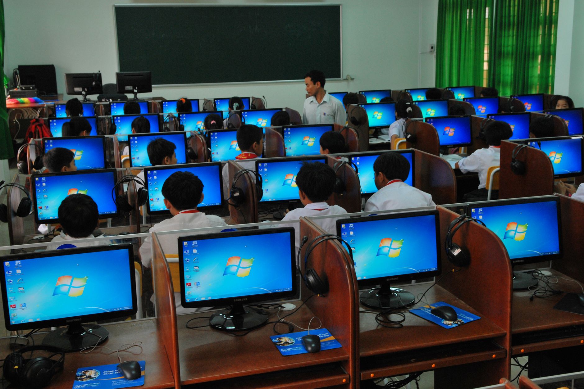 Students learning in a computer lab with multiple monitors and a teacher instructing — UPI Study