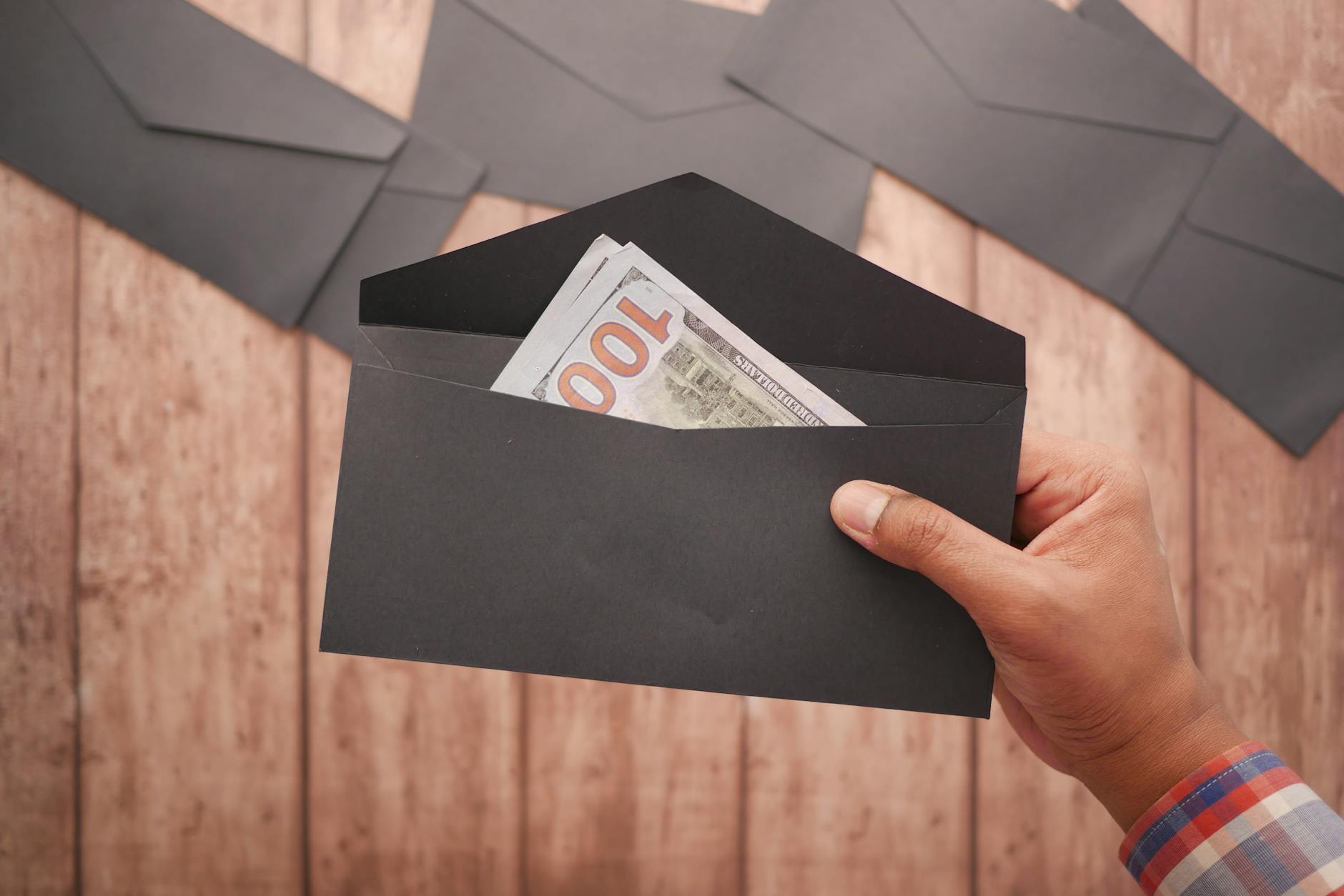 A hand holds a black envelope containing a 100 dollar bill on a wooden surface — UPI Study