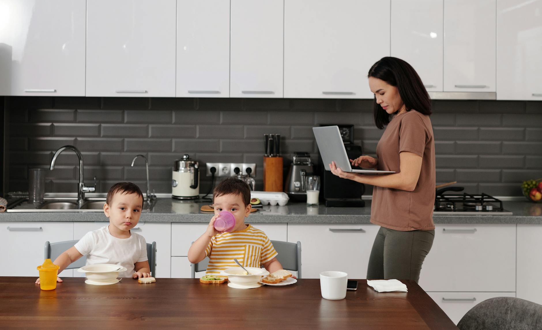 A mother multitasking with a laptop while her children enjoy breakfast at home — UPI Study