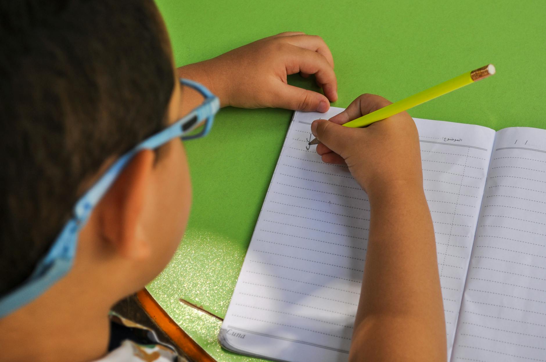 A child wearing glasses writes in a lined notebook with a yellow pencil on a green table — UPI Study