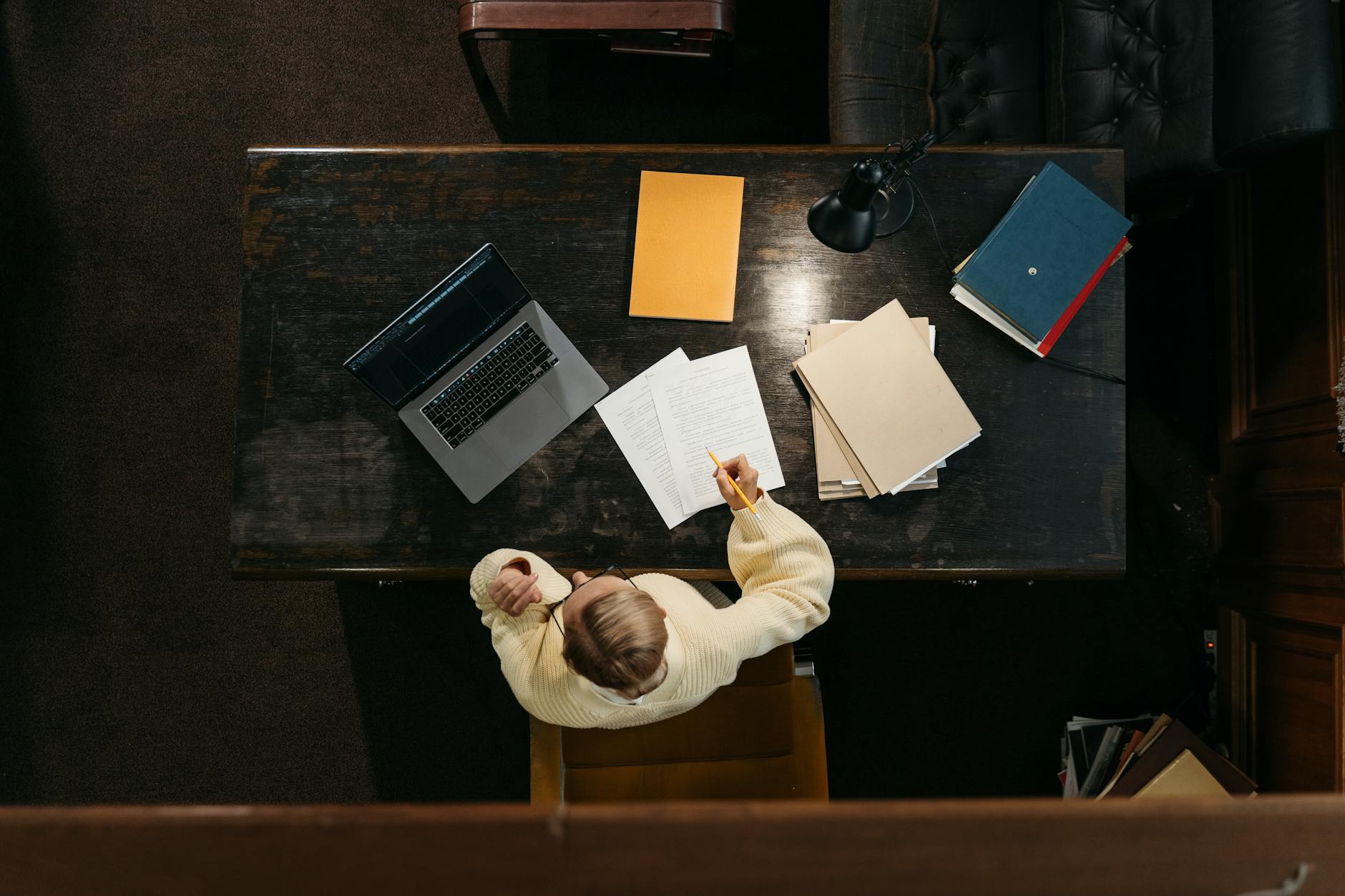Person in yellow sweater working at a wooden desk with documents, folders, and a laptop — UPI Study